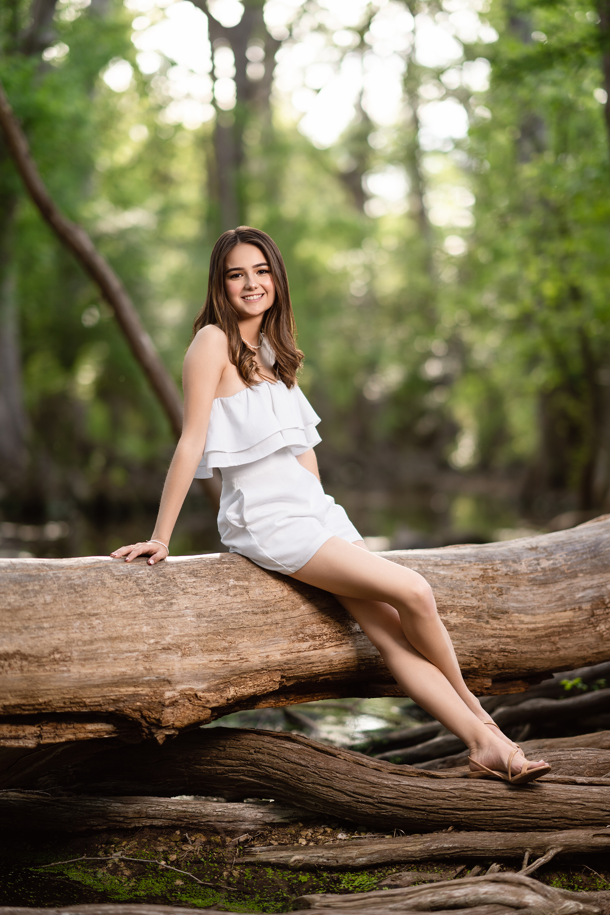 Relaxed outdoor portrait of a young woman in a white summer outfit seated on a fallen tree in a wooded setting