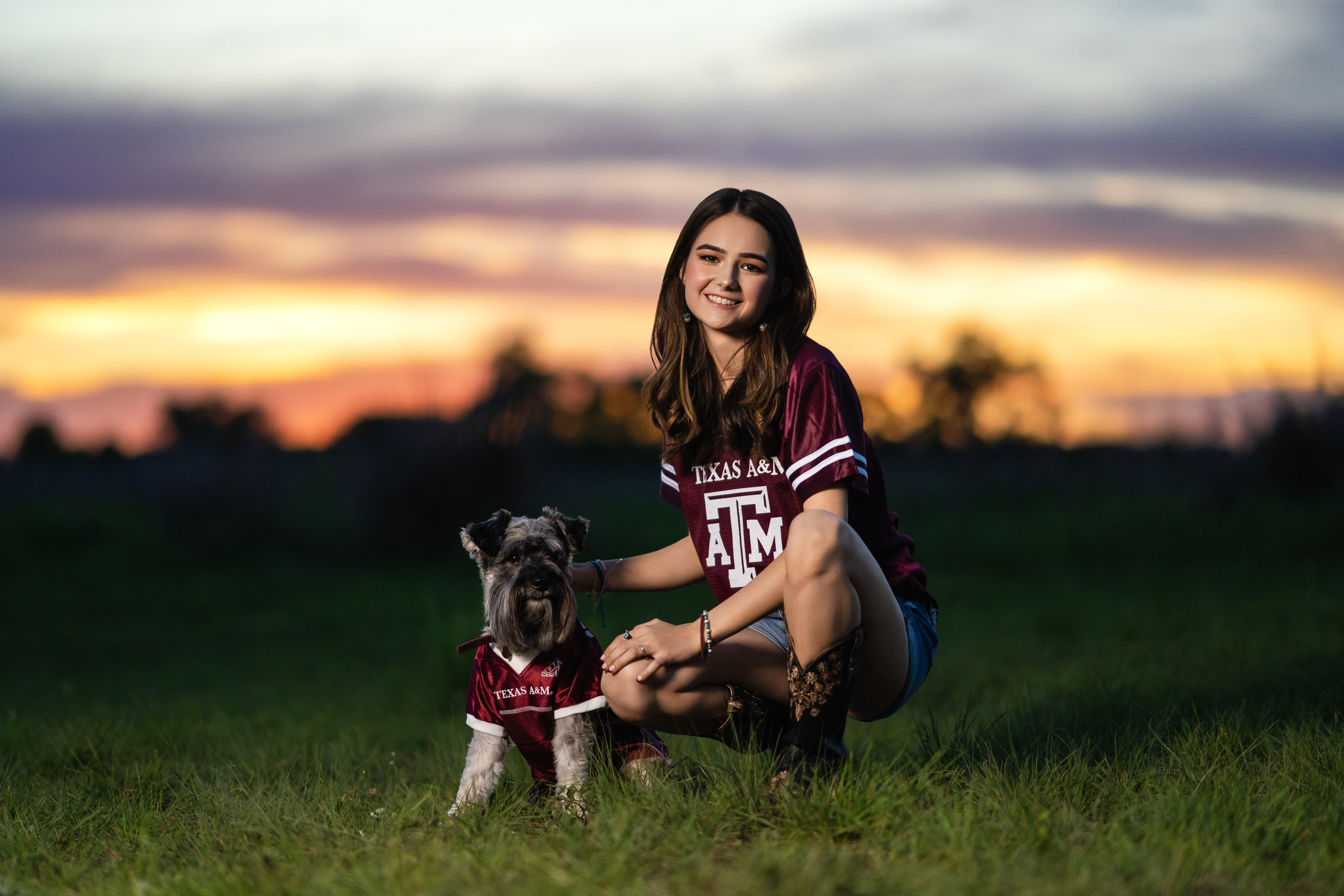 Sunset senior portrait of a young woman in a maroon college shirt seated in a field with her dog