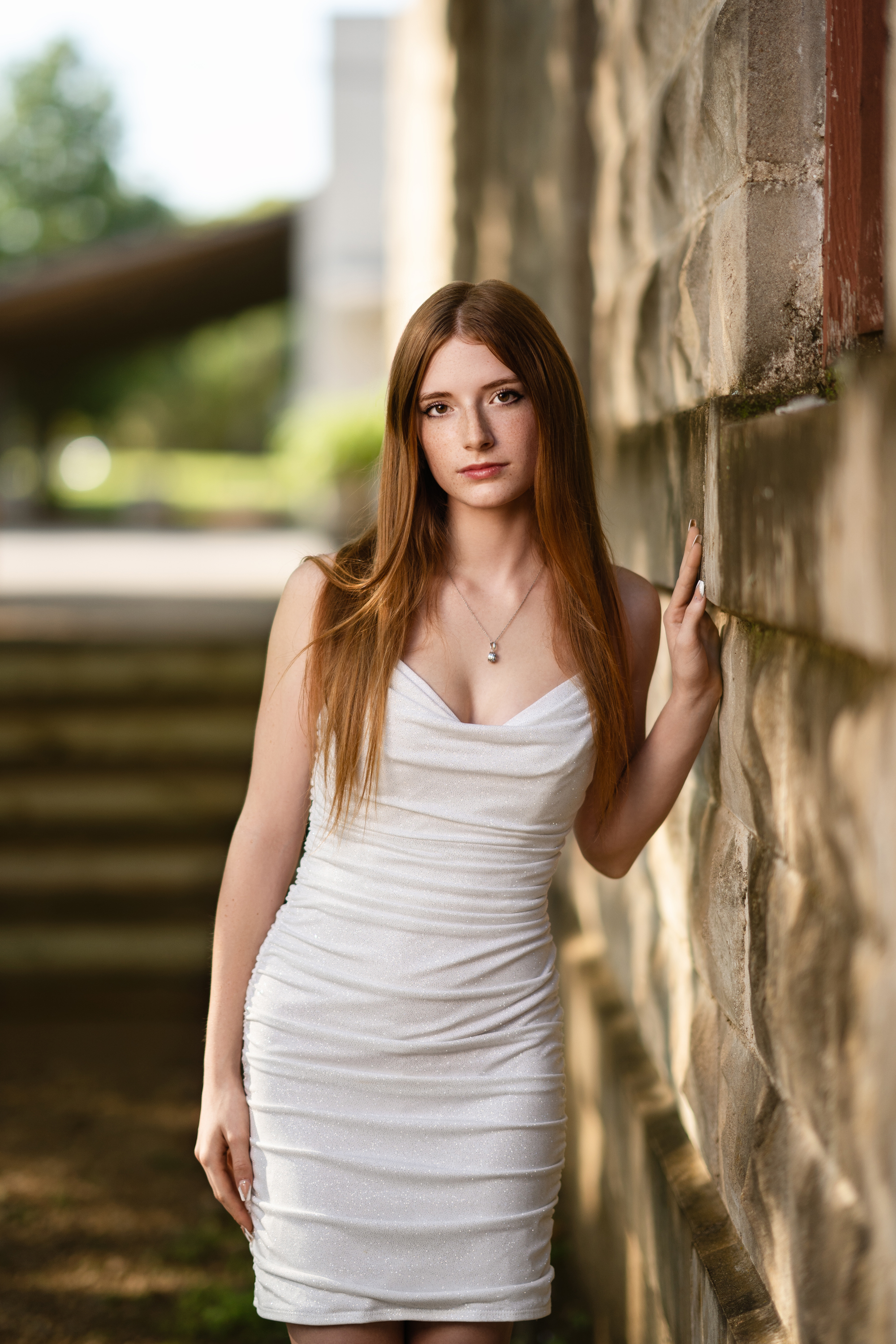 Outdoor graduation portrait of a young woman in a white dress standing beside a rustic stone wall