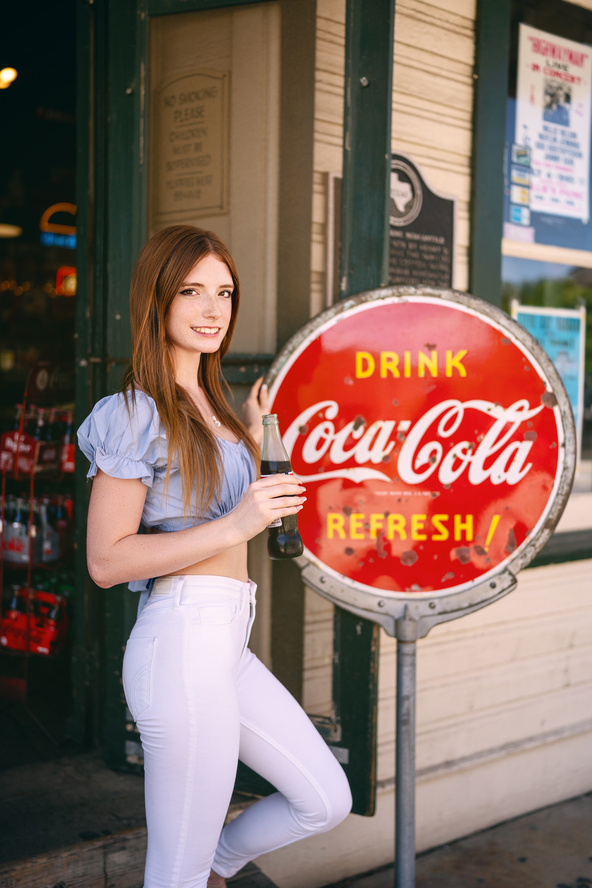 Lifestyle portrait of a young woman in a pale blue top posing beside a vintage Coca-Cola sign