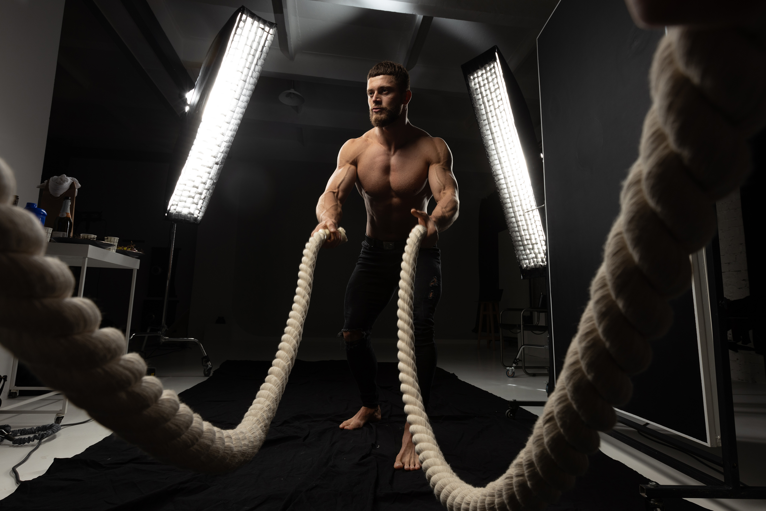 Behind-the-scenes fitness image of a shirtless man working with battle ropes in a studio before retouching