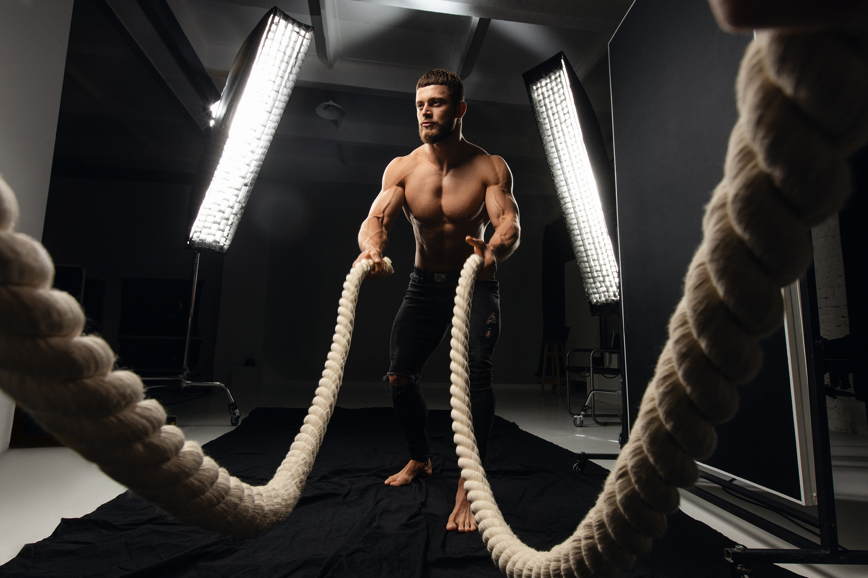 Wide athletic portrait of a shirtless man training with battle ropes under dramatic studio lighting