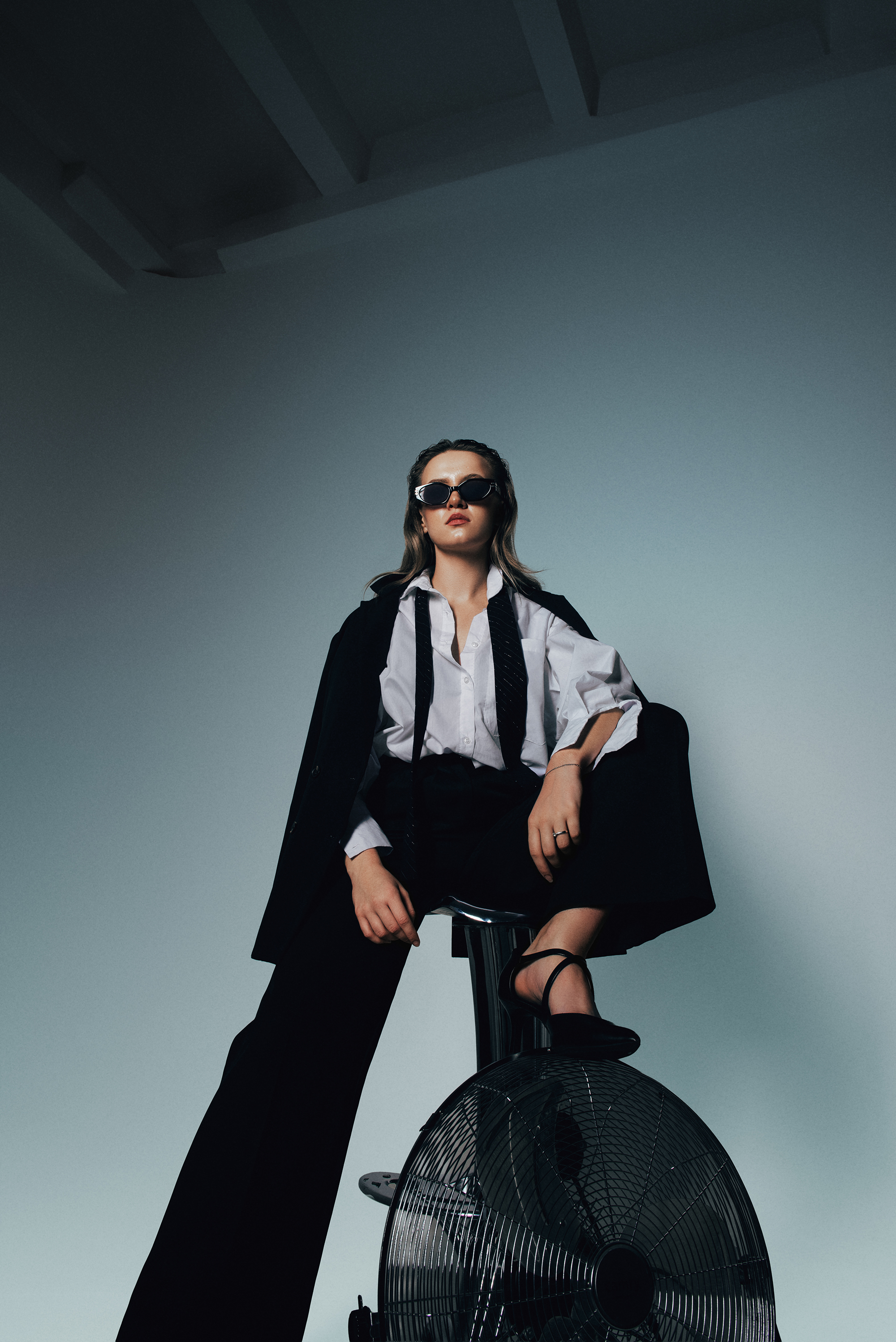 Fashion portrait of a woman in sunglasses and black-and-white tailoring seated above a large floor fan against a cool blue-gray background