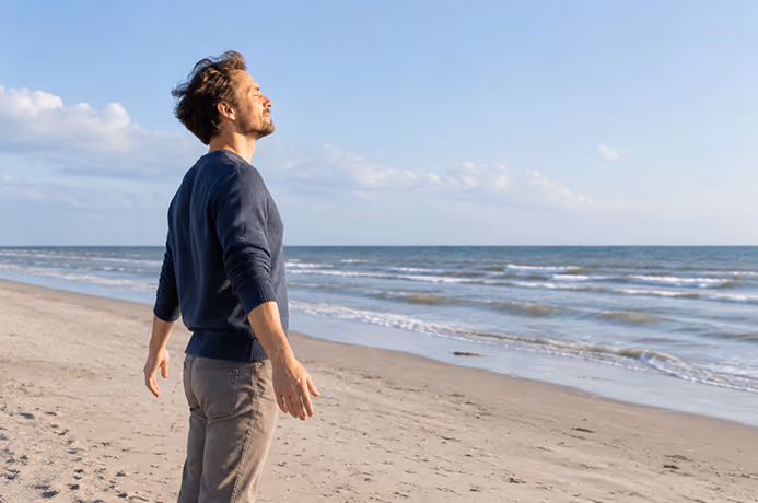 Mann atmet tief ein, steht mit geschlossenen Augen entspannt am Sandstrand mit Blick aufs Meer.