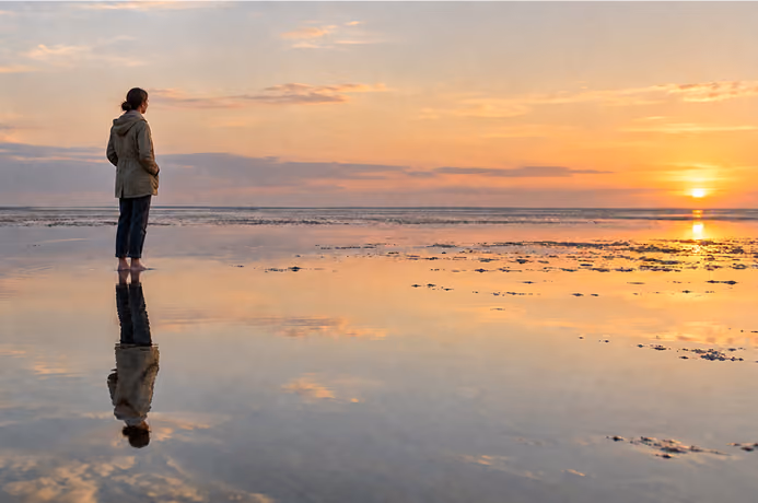 Eine Frau steht barfuß im flachen Wasser in der Nordsee und schaut Richtung Sonnenuntergang.