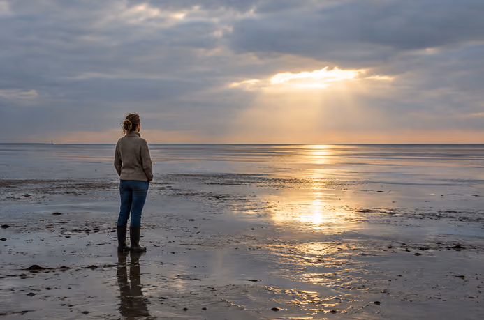 Eine Frau steht im Watt und schaut bei bewölkten Himmel weit auf die Nordsee hinaus.