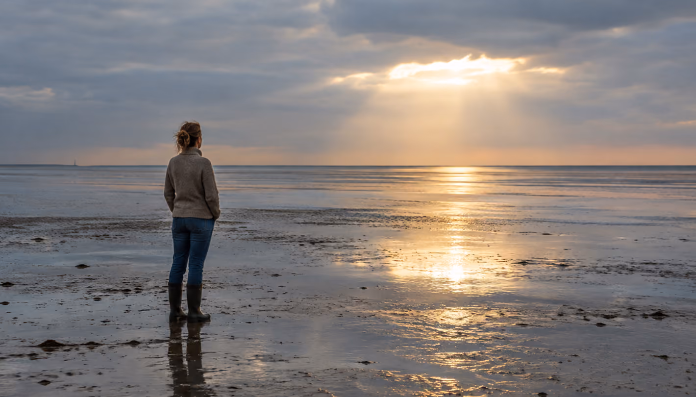 Eine Frau steht im Watt und schaut bei bewölkten Himmel weit auf die Nordsee hinaus.