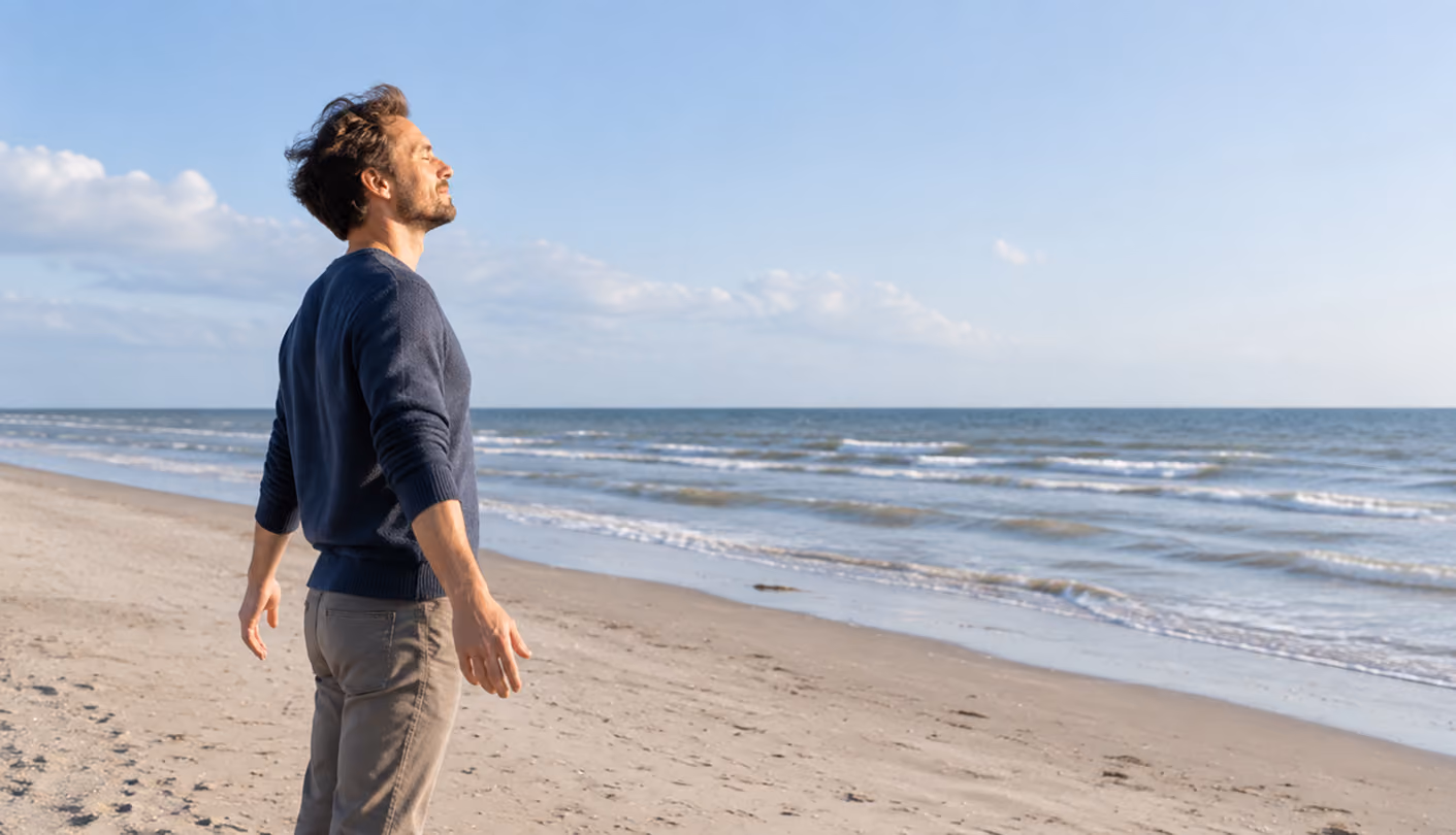 Mann atmet tief ein, steht mit geschlossenen Augen entspannt am Sandstrand mit Blick aufs Meer.