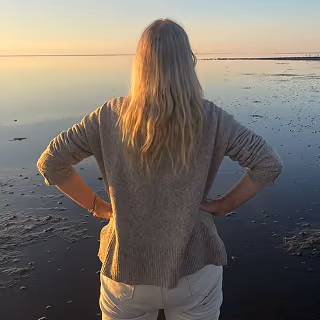 Emma Köpnick steht am Ufer eines ruhigen Gewässers steht und den Sonnenuntergang betrachtet.
