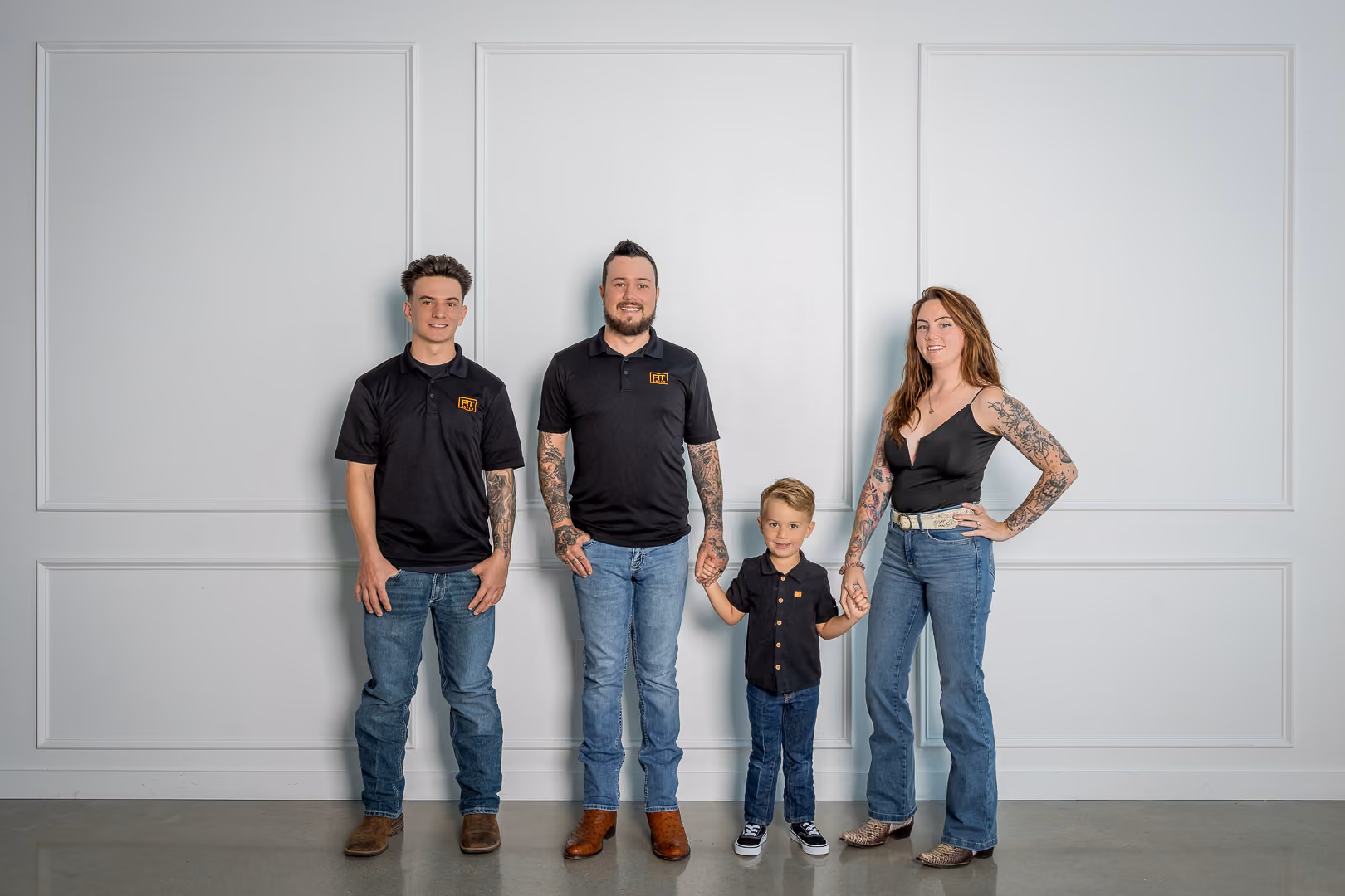 Family of four holding hands, dressed in black tops and blue jeans, standing against a white wall.