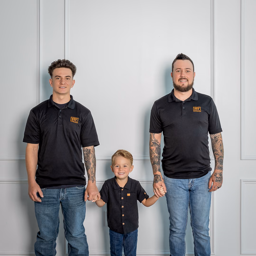 Two men with tattoos and a young boy holding hands, all wearing black shirts and jeans, smiling.
