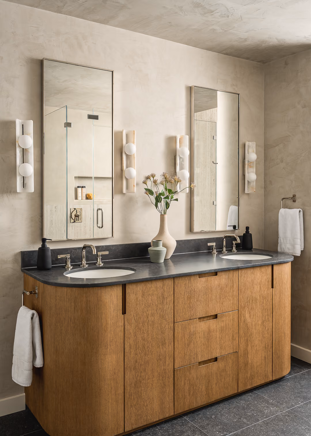 Modern bathroom with double sink vanity, wood cabinet, two mirrors, wall sconces, and a vase with flowers.