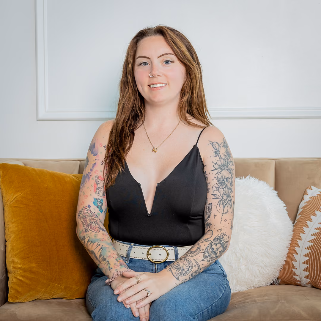 Smiling woman with long brown hair and arm tattoos sitting on couch with decorative pillows.