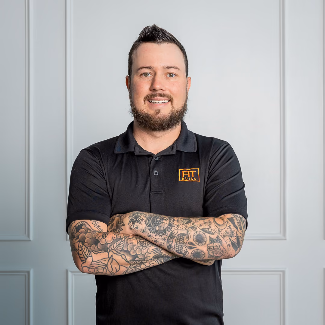 Man with beard and tattoos wearing a black Polo shirt with FIT BUILD logo, arms crossed, smiling.
