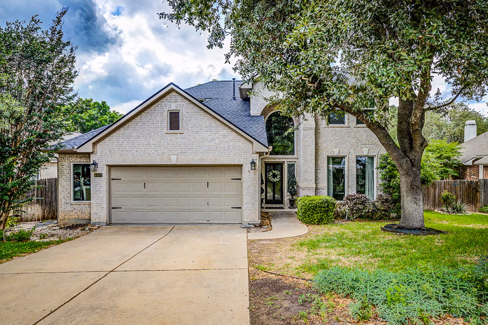Two-story brick house with gray roof, double garage, large front tree, and green lawn.