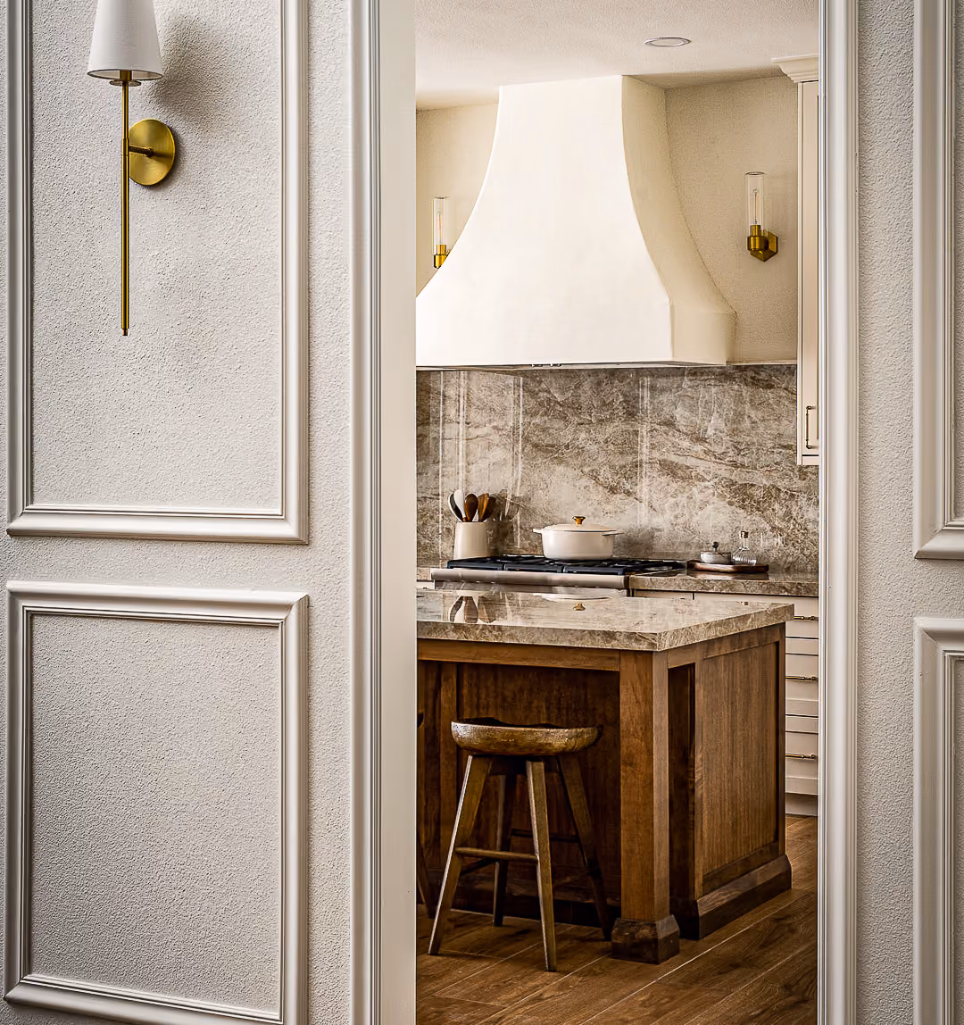 View through doorway into kitchen with marble island, wooden stool, stove, and white range hood.