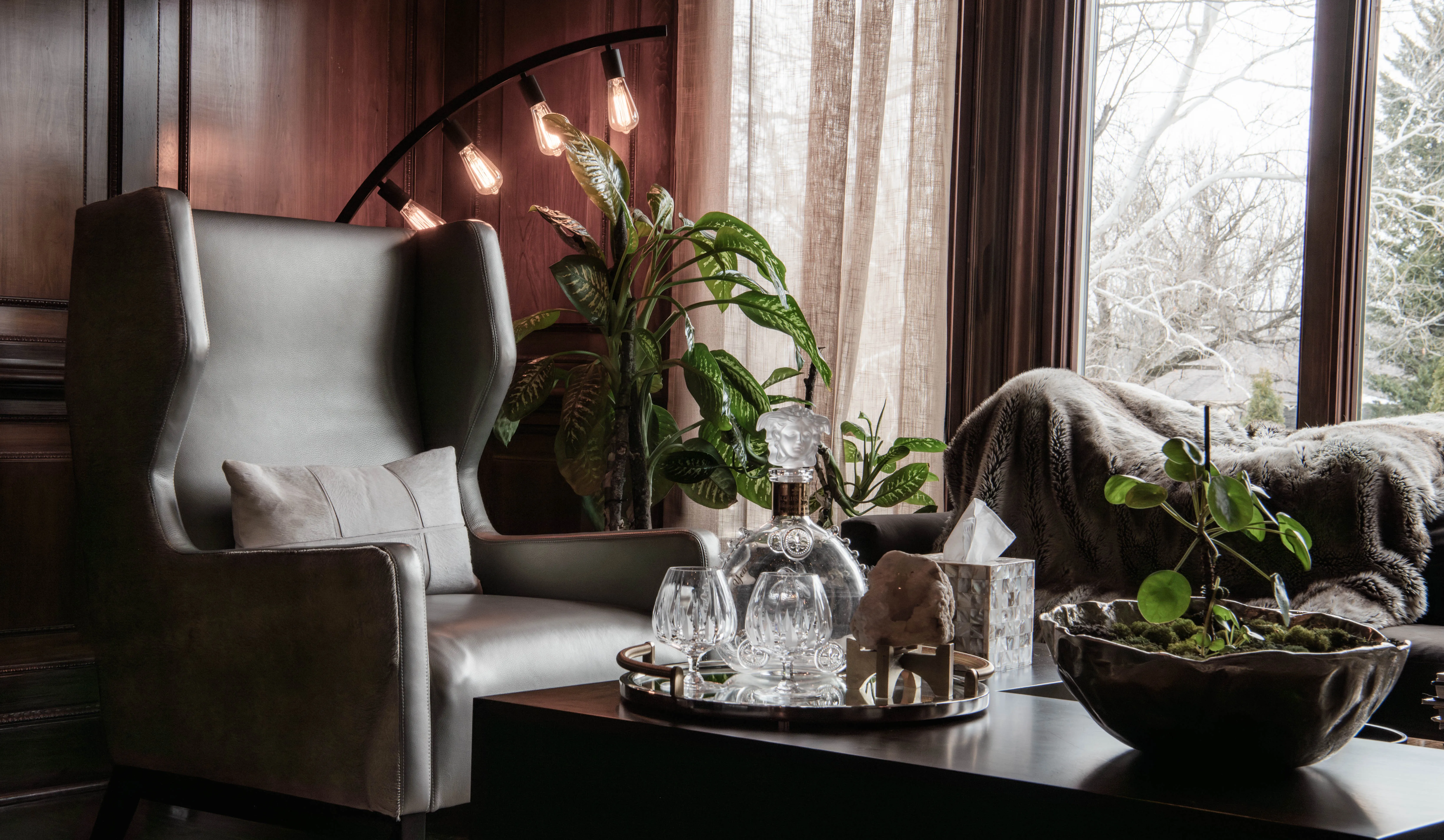 Luxury modern home interior with a gray leather wingback chair; the sides are lined with real animal hide. A modern floor lamp sits next to the chair, with Edison bulbs and tropical plants.
