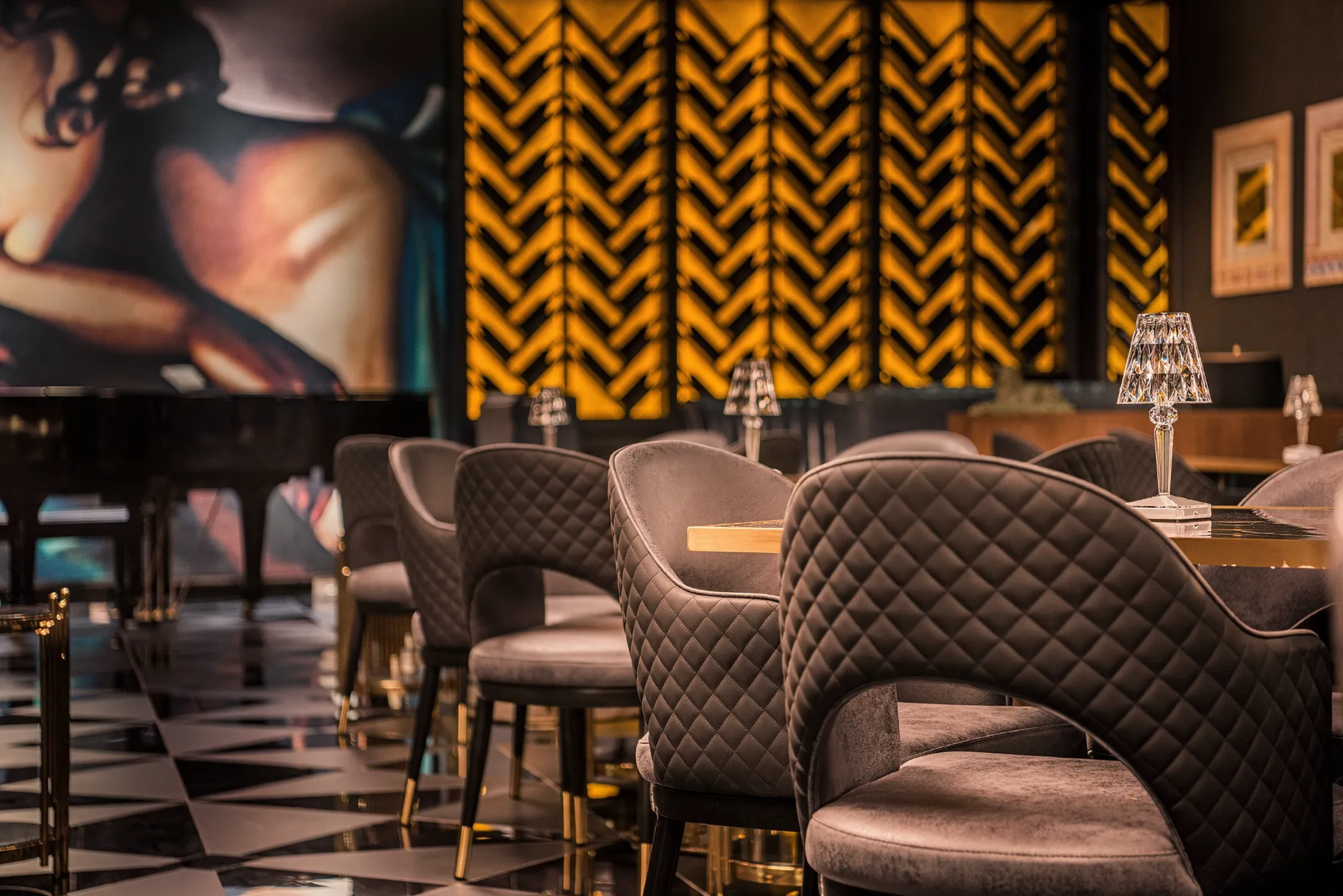 Art Deco luxury interior, showing custom-made Bentley-style leather stitching and multi-finish black floor. The tables are marble tops with gold frames and crystal table lamps. The background is an Art Deco full-wall-height artwork, beside oversized wine display wall units, backed with backlit onyx. 