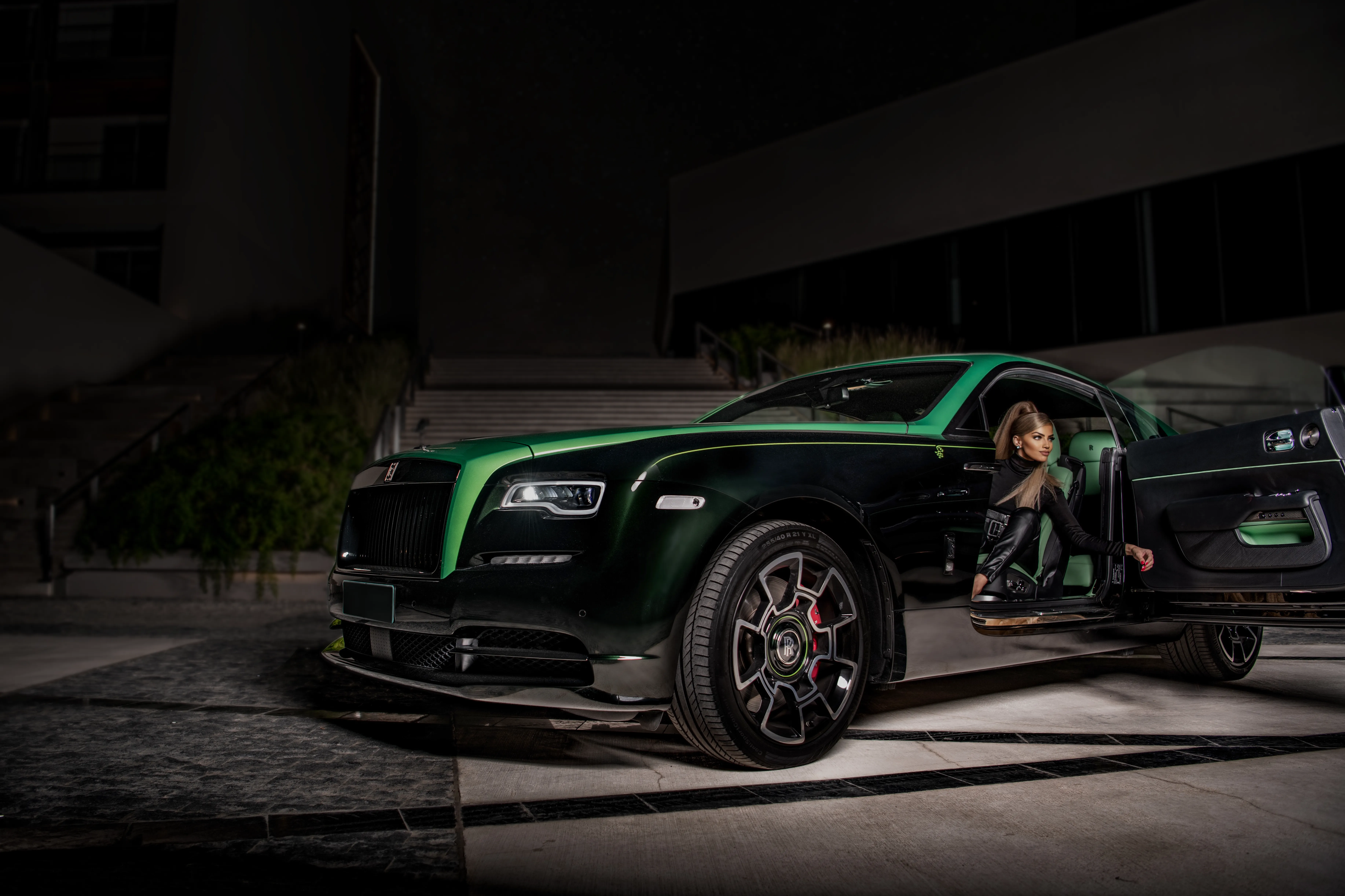 Black and green Rolls-Royce Ghost, with the model opening the door, wearing leather pants and a dark jacket. The background is an urban structure with the night sky behind it.  