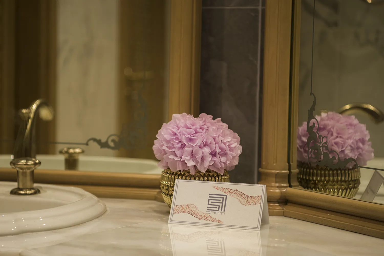 A pink hydrangea flower in a luxury bathroom with a decorated mirror in the background and SANTI Design House’s calling card in front of it.