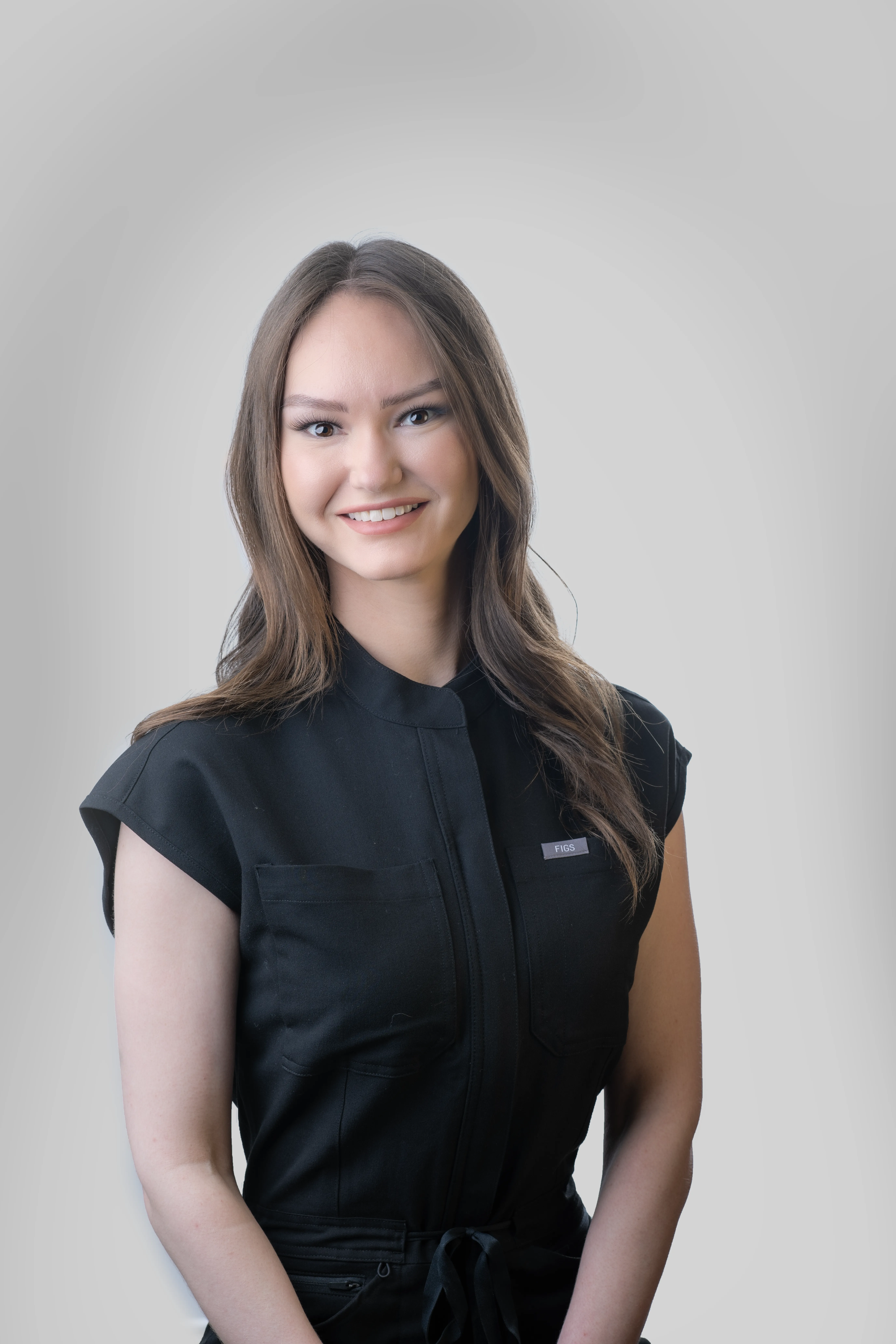 Professional headshot for medical staff, with dark brown hair on a white background. Wearing dark gray scrubs and smiling. In portrait format. 