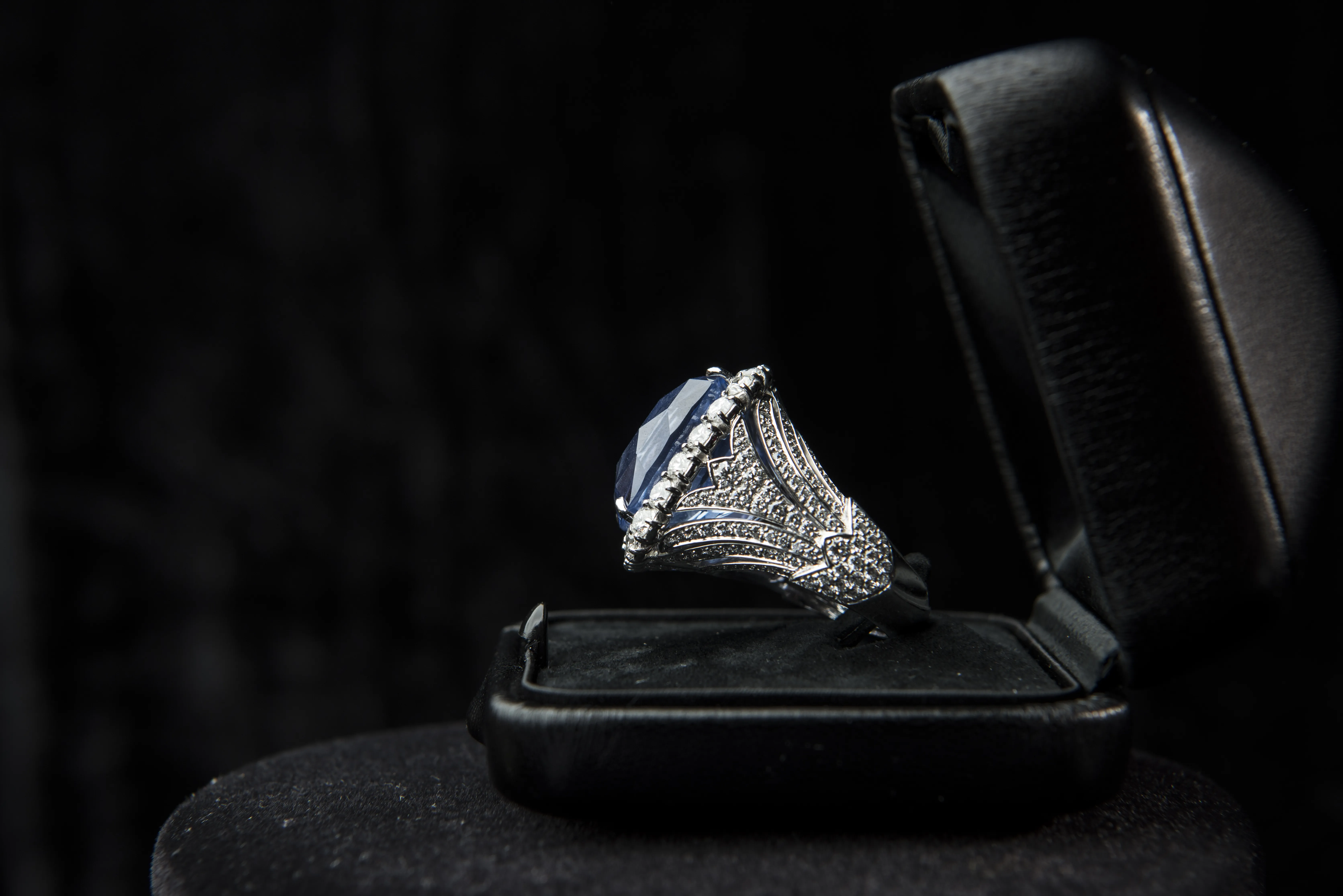 A side profile of a luxury ring, made of white gold and a large blue stone, sits on a velvet and leather ring box. Detailed metalwork. 