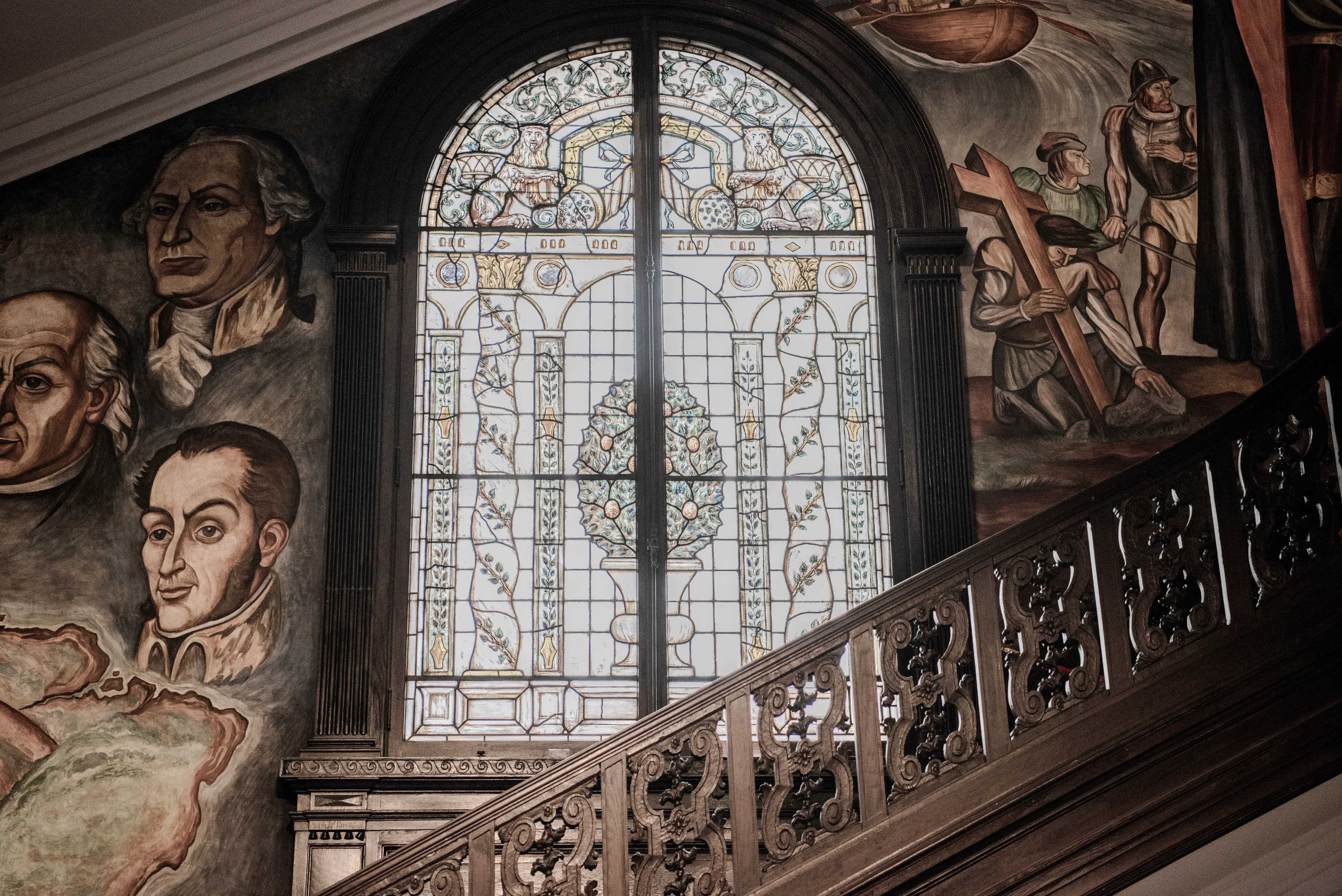 Traditional staircase with carved wood and a mural in the background. A large arched stained glass window in the middle. 