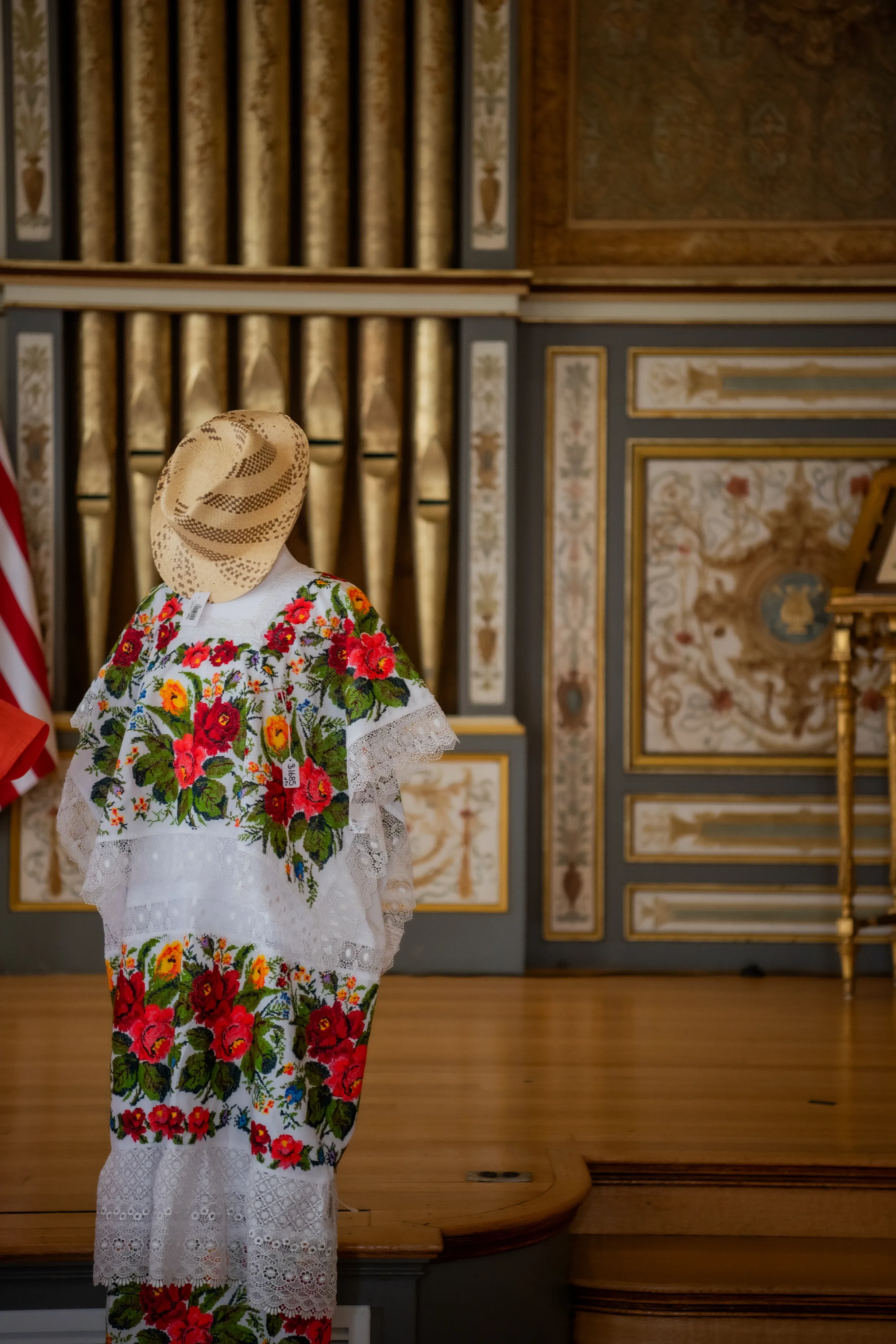 Architectural shot of an ornate interior, with traditional imperial Spanish design. With large gold organ pipes and a traditional dress in the foreground, and a flag to the side. 