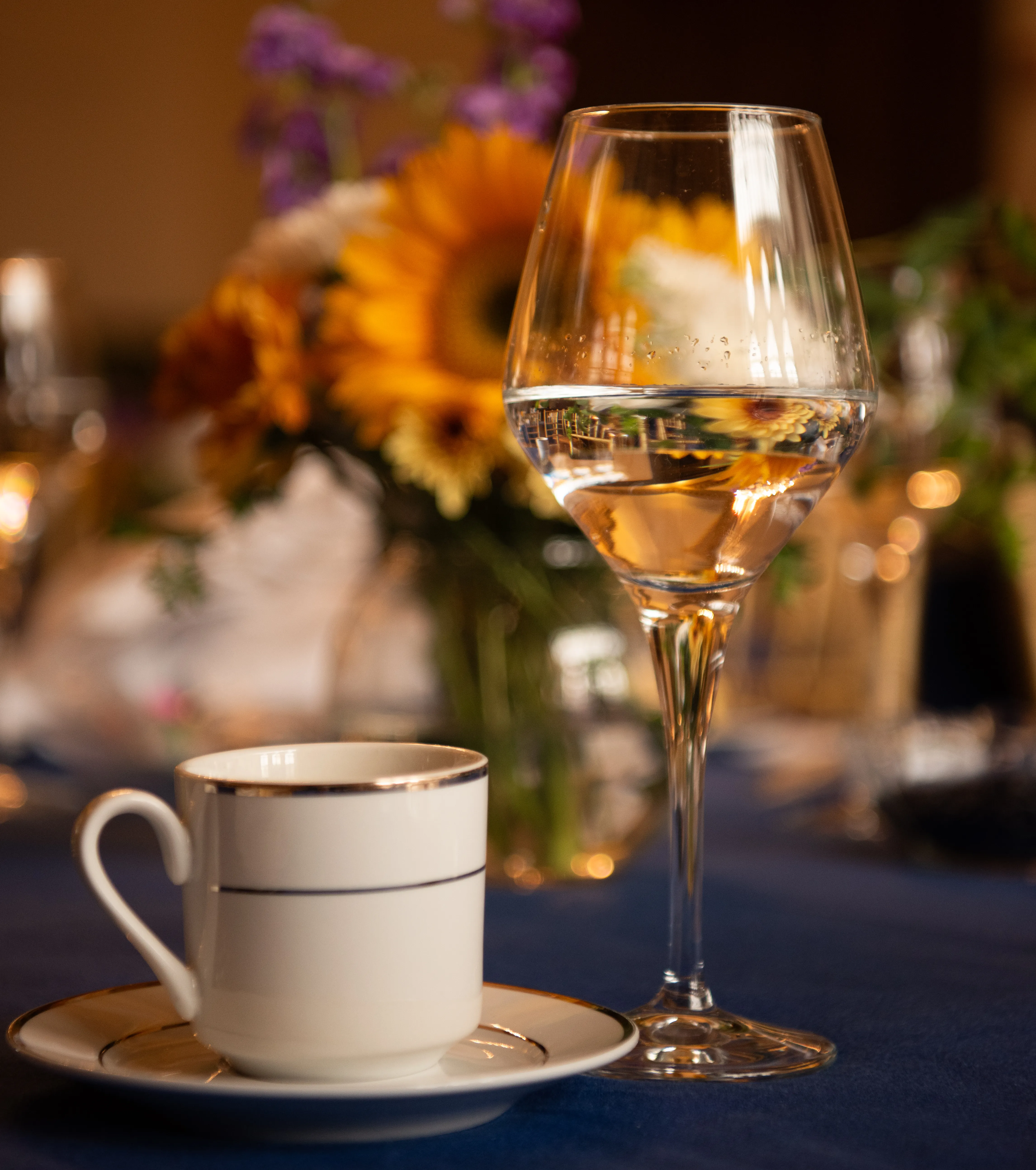 Close-up shot of a white and gold coffee cup and a white wine glass. With flower arrangements blurred in the background, on a dark blue tablecloth.