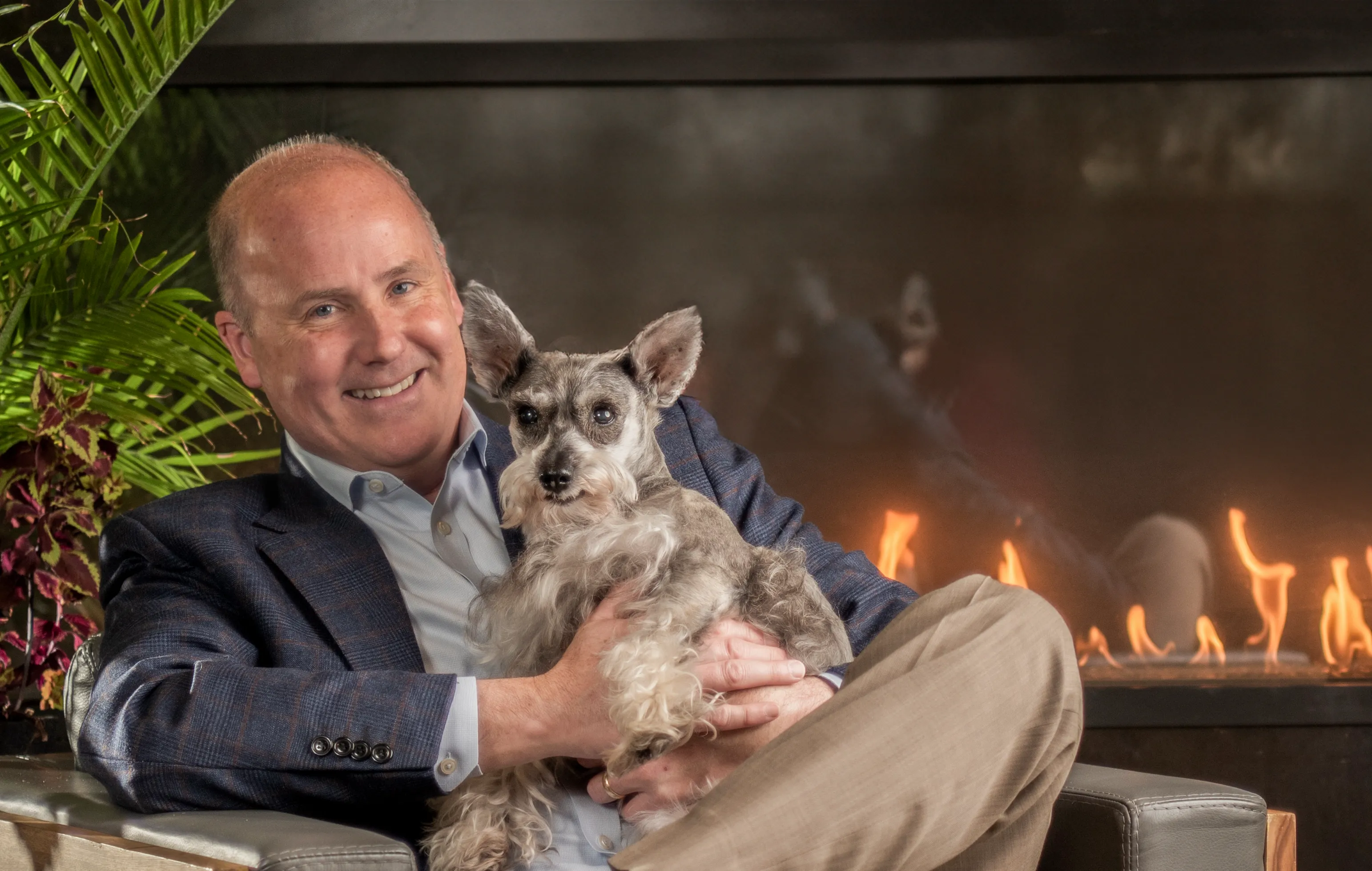 Family photograph of the father holding the family pet, wearing a casual suit with brown khakis. Next to a tropical plant, with a modern oversized fireplace in the background. 