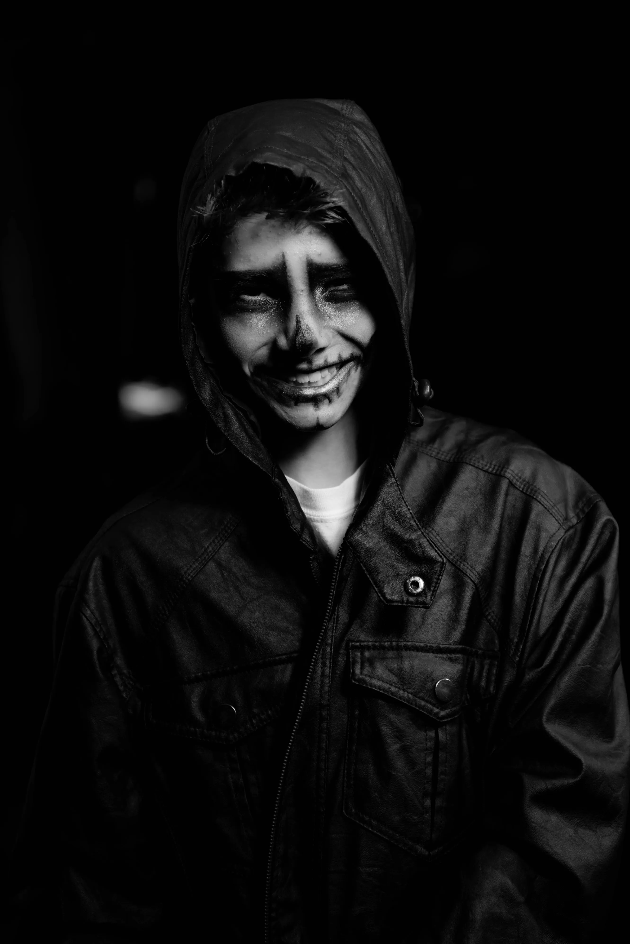 Black and white photograph of a child in Halloween makeup smiling. Wearing a leather jacket. Heavy contrast and black background. 