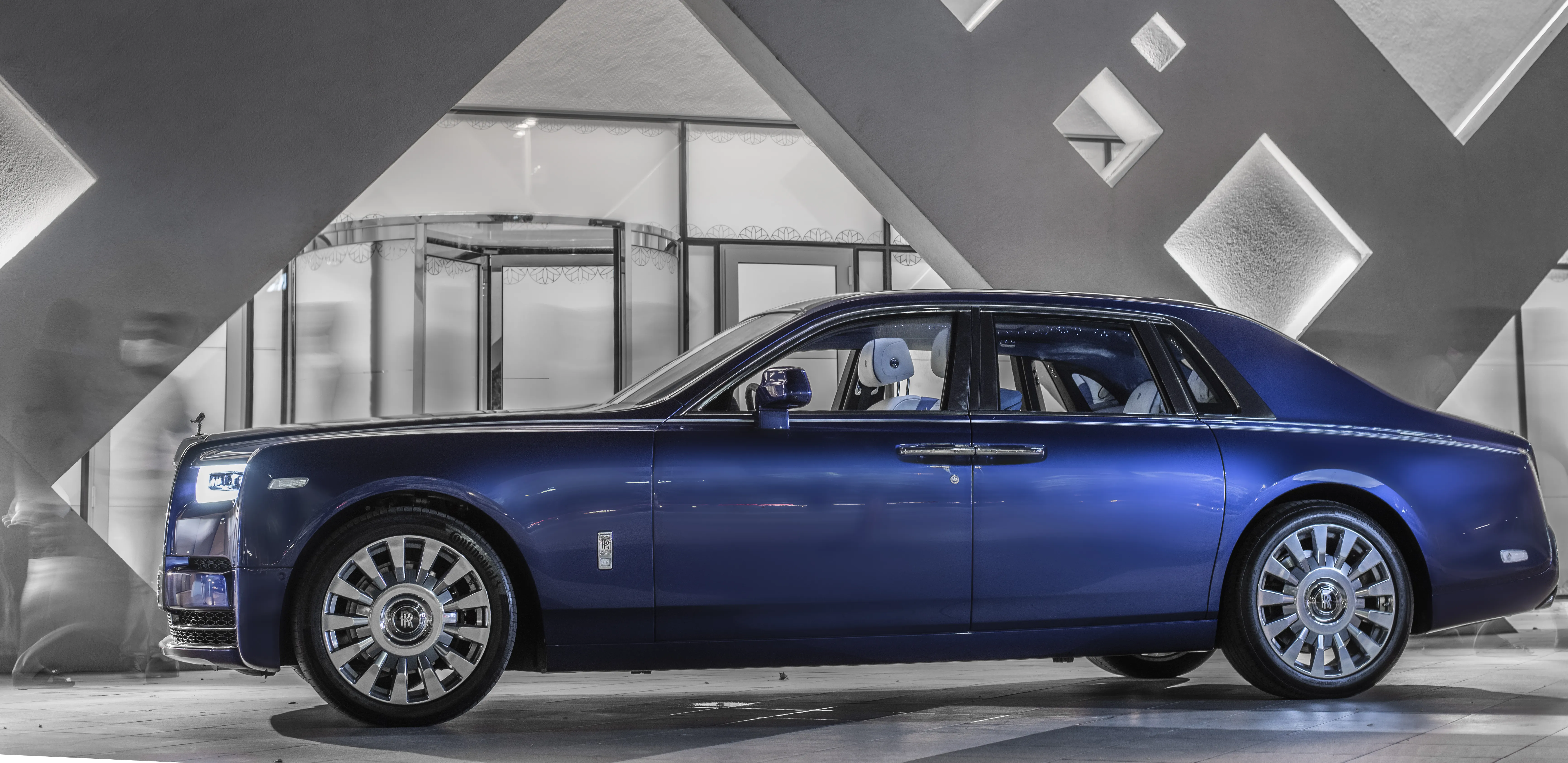side profile of a blue Rolls-Royce Phantom photographed in front of modern fractal architecture . People fading in the background through long exposure. Monochromatic photograph.