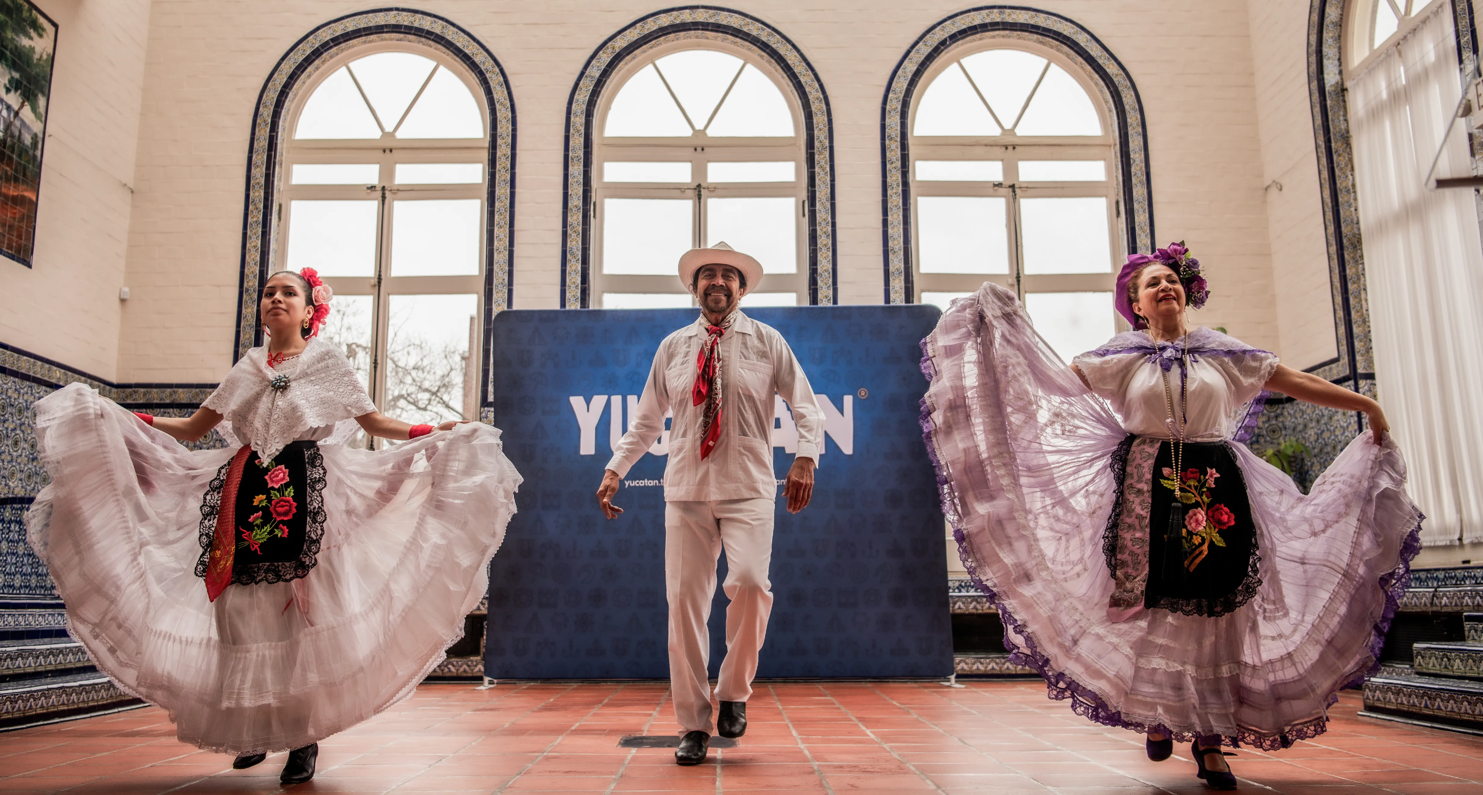 Cultural event, with 3 traditional Mexican dancers taking the stage. Day event, with large arched windows in the background and a blue event banner. 