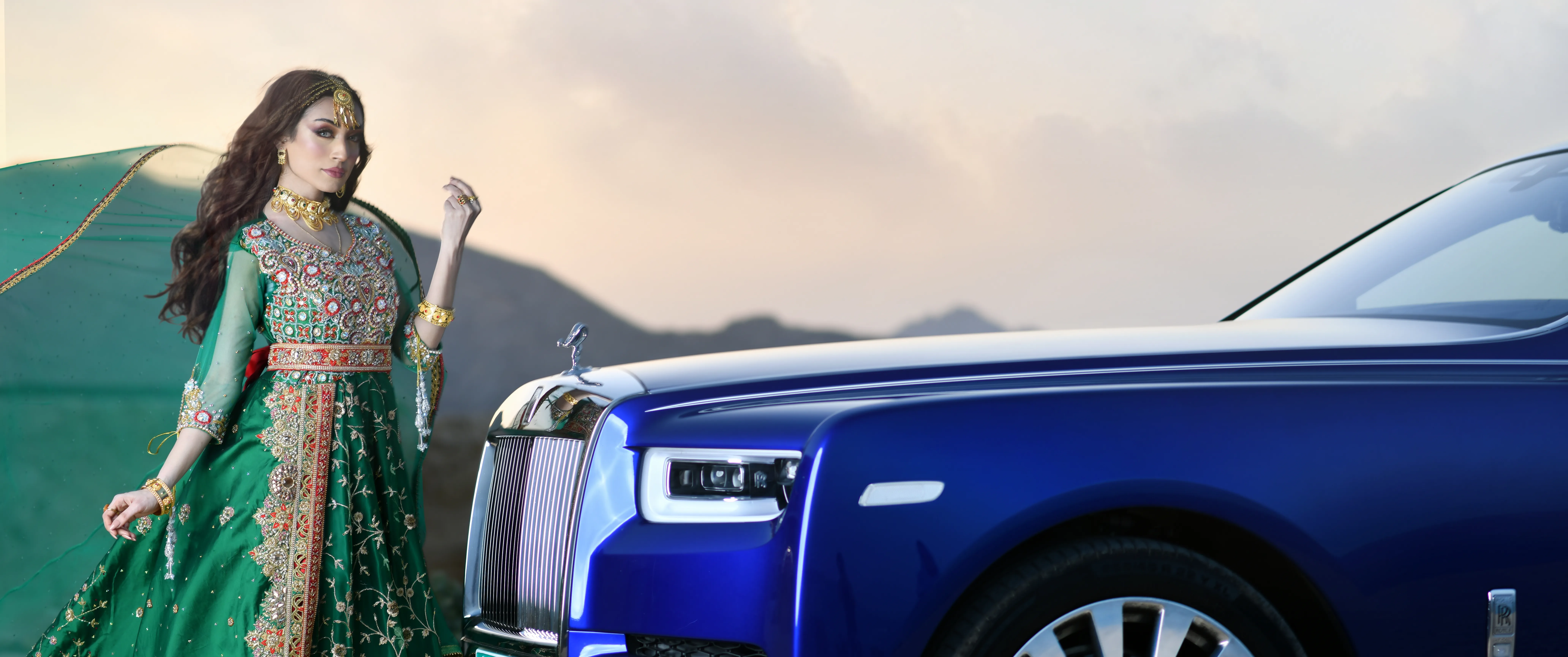 A model in a green ornamental dress standing next to a blue Rolls-Royce Phantom at sunrise. The location is a cliff with mountains in the background under the orange sky. 