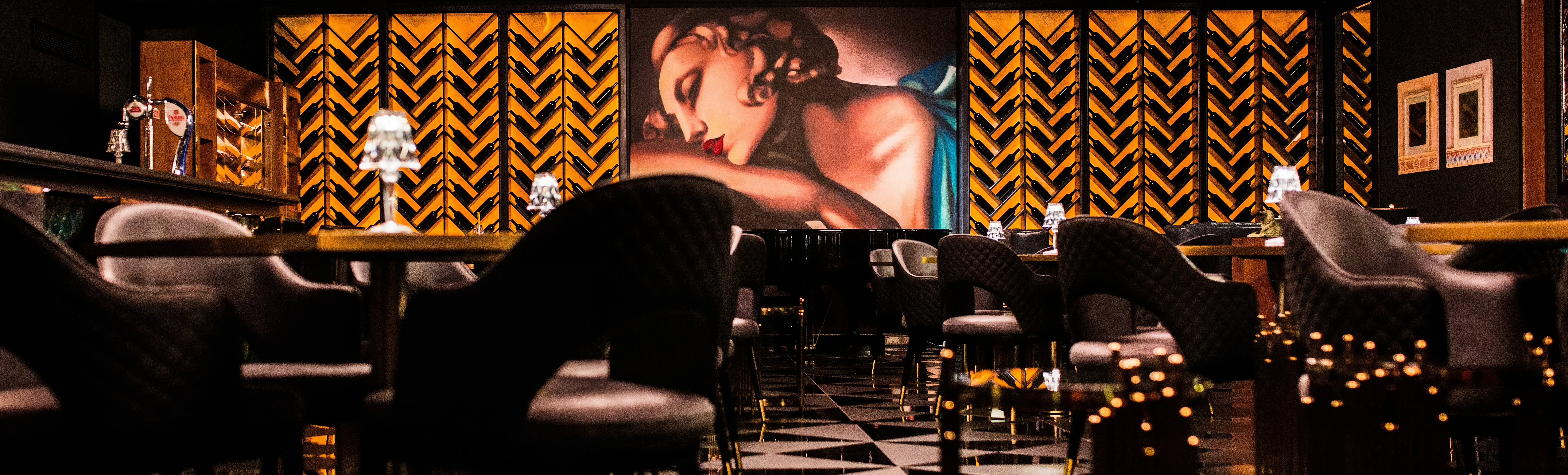 Luxury cigar lounge and bar, with leather Bentley-style chairs, a custom multi-finish black floor, and gold and black glass tables. At the center is custom large-scale Art Deco artwork of a woman with natural tones and a turquoise dress. Flanked by wine display angled shelves on lit onyx.  