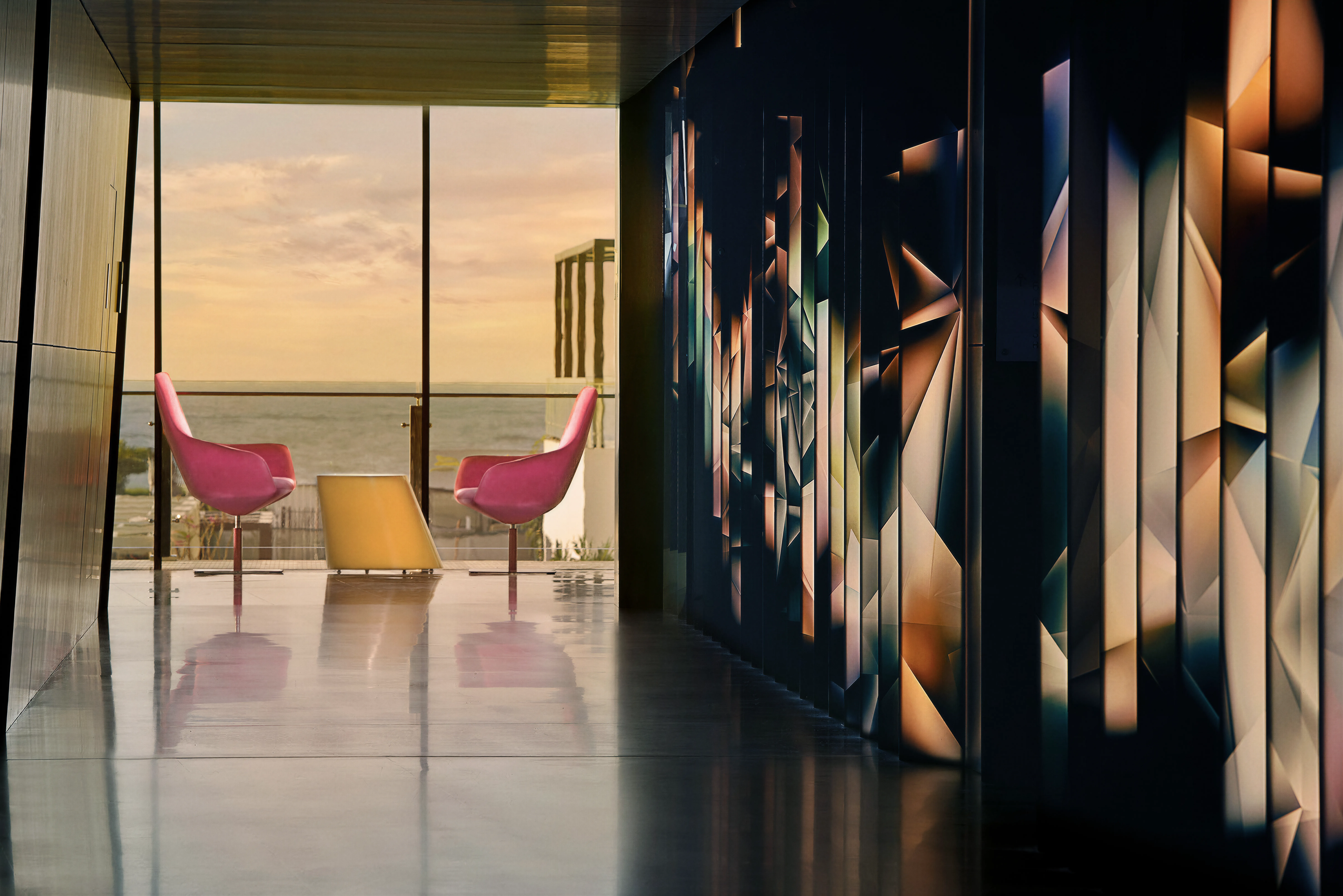 Hotel hall at sunset, featuring dtop-tix fractal backlit artwork, placed at a 45-degree angle. The background is the ocean at sunset, with 2 pink chairs and a yellow coffee table. 