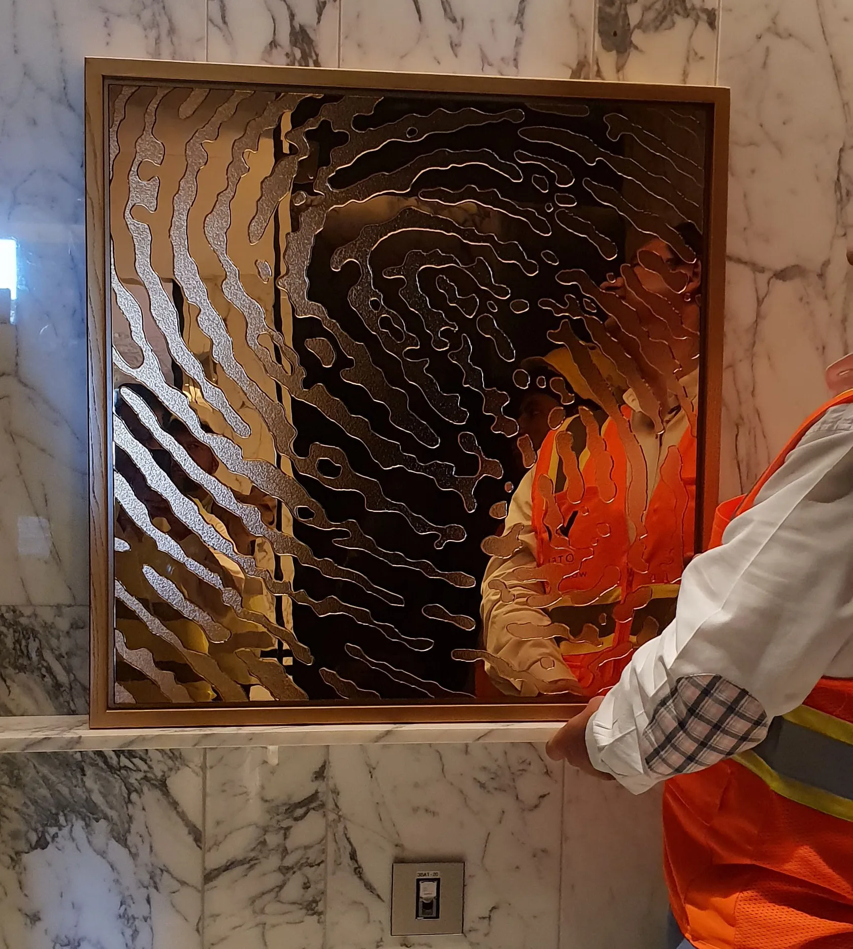 Golden glass fingerprint 3D glass artwork on a golden frame, being hung by a worker in a hotel bathroom. White marble wall.