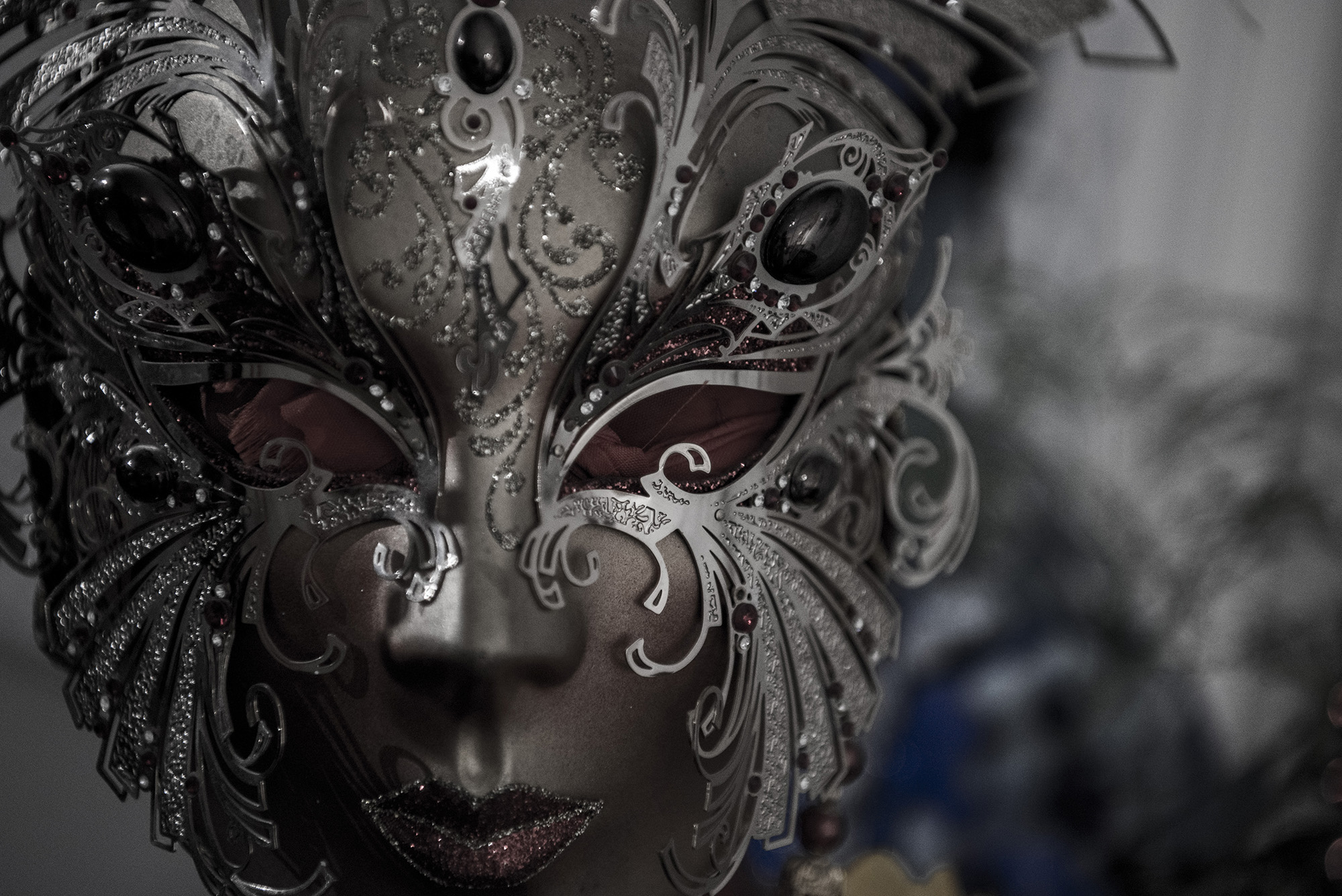Close-up shot of a Venetian mask, placed on a mannequin, with an artistic background. The mask is silver with red stones and intricate metalwork.