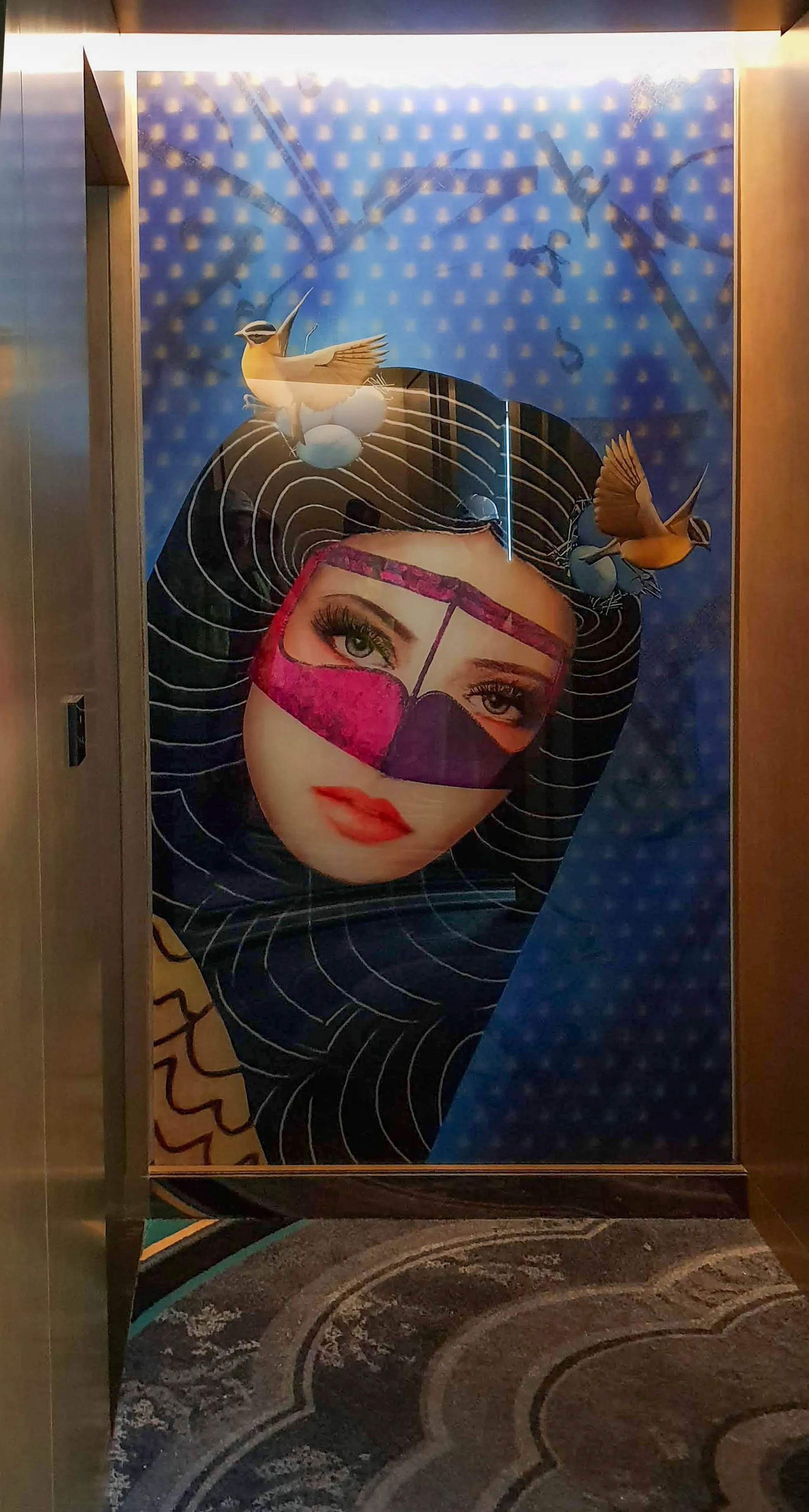 Lenticular wall art in a hotel corridor features interactive appropriation art, with the mask fading as you walk by. Birds move against a blue background with black hair.
