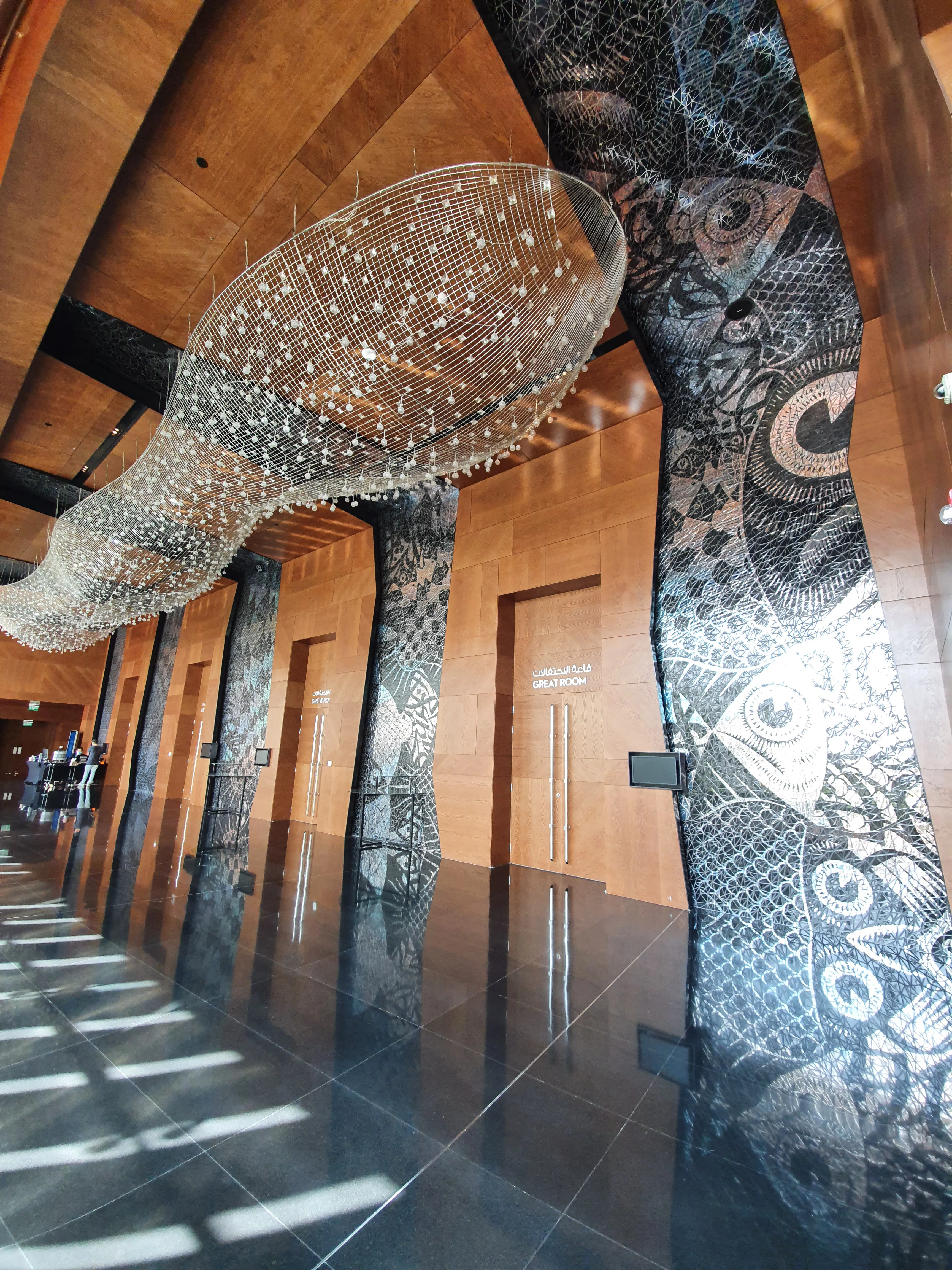A resort ballroom main hall, featuring large-scale appropriation art and a custom ceiling. Wood cladding and recessed lighting. Dark stone floor. Morning light casts light on the artwork.  