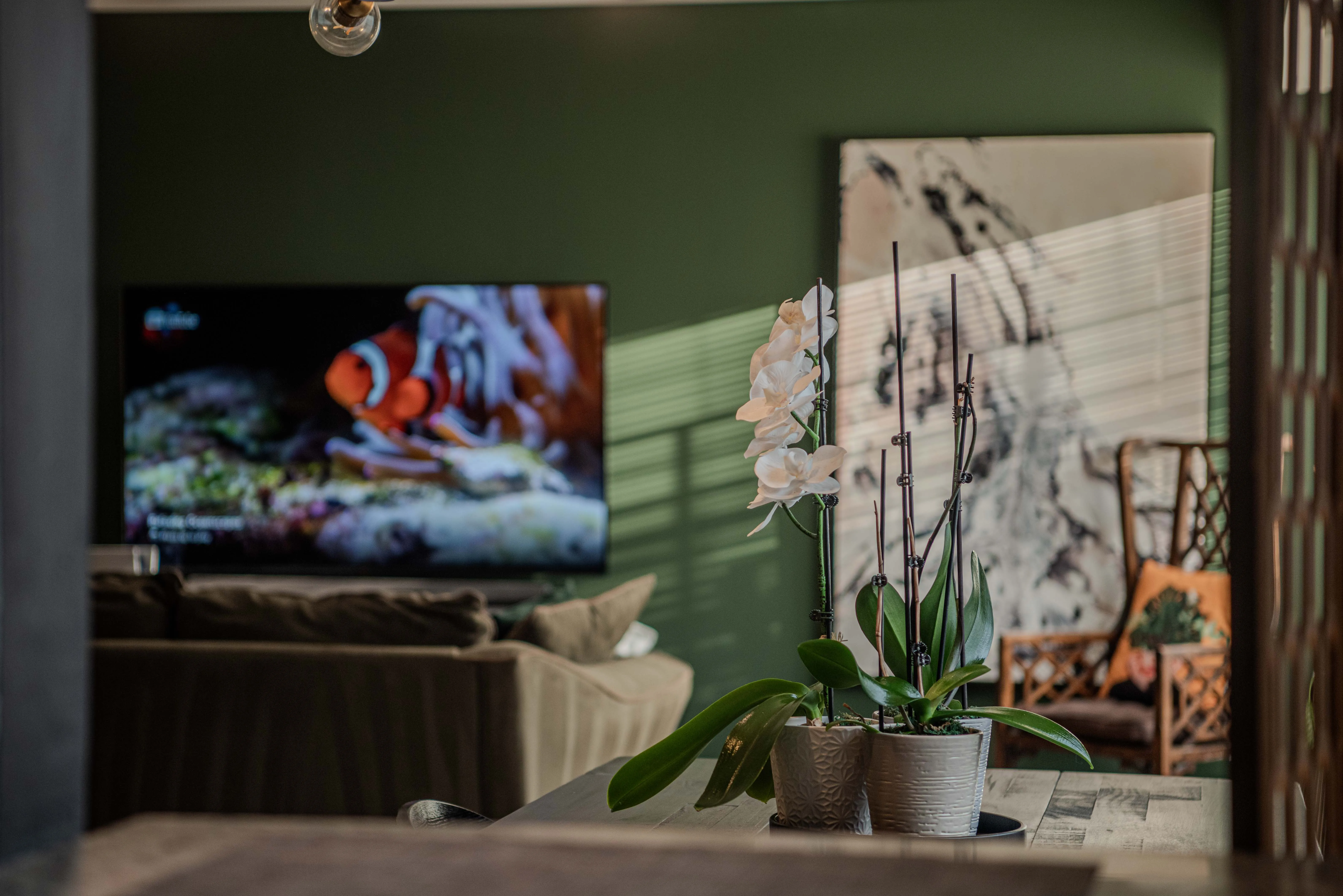 Modern home staging with an olive green sofa and custom pillows, a deep green feature wall, a white orchid flower on the dining table, a wooden screen, and a modern white and deep blue painting.