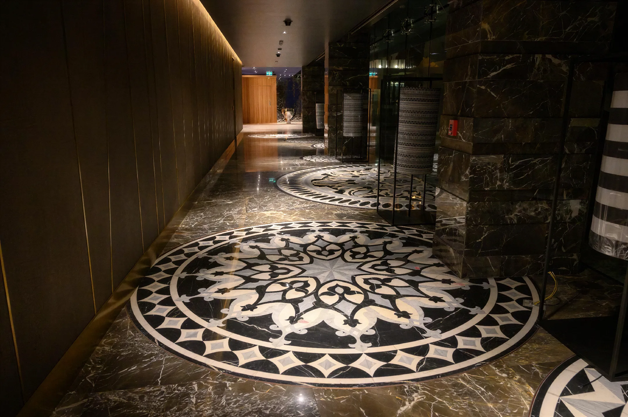 Hotel interior, highlighting custom natural stone medallion flooring, in black and white. On the right, polished black marble and glass walls, with custom floor lamps. And on the left, canvas walls with gold trim.