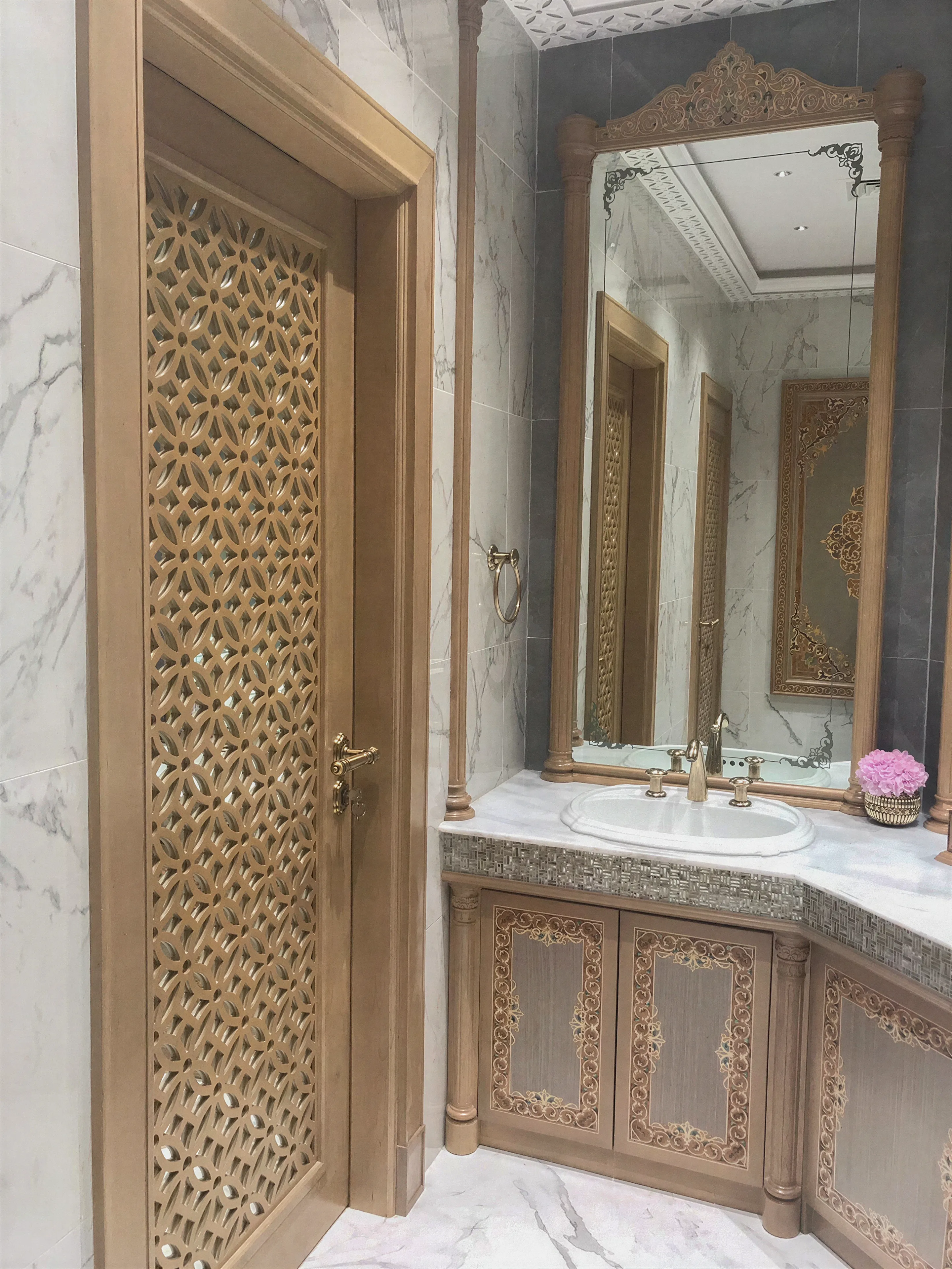Door view of a luxury bathroom with a custom inlay cabinet, mother-of-pearl counter edging, and a white porcelain countertop. Ceiling-high decorated mirror in a custom beachwood frame. Dark gray granite walls and custom-milled beachwood doors. 