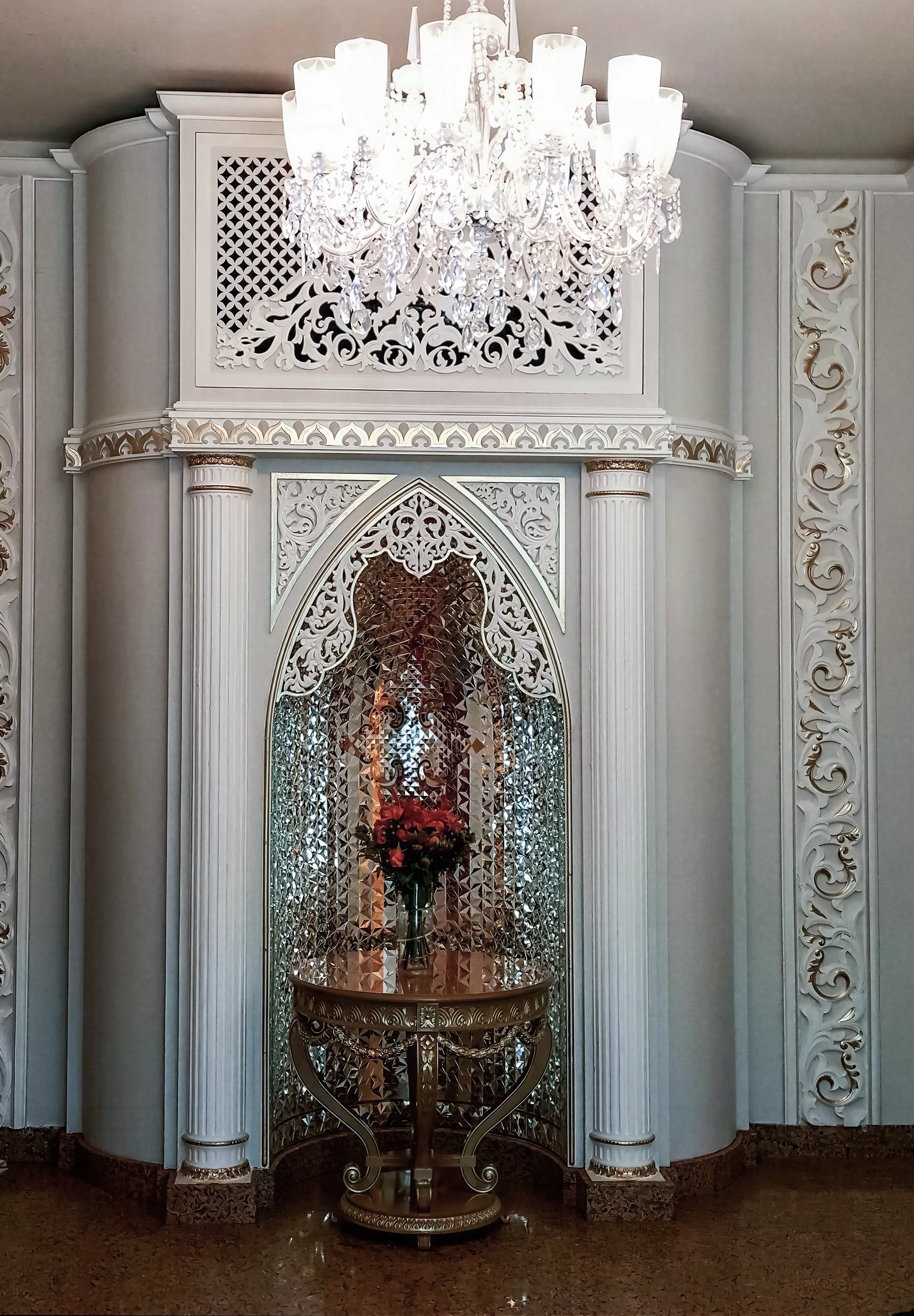Classic luxury home with a cathedral-vaulted niche, a fractal glass interior, and a white-and-gold exterior. A custom-made wooden entryway table and a flower arrangement are placed in the center of it.  A crystal chandelier hangs from the ceiling, and the floor is dark, polished granite.