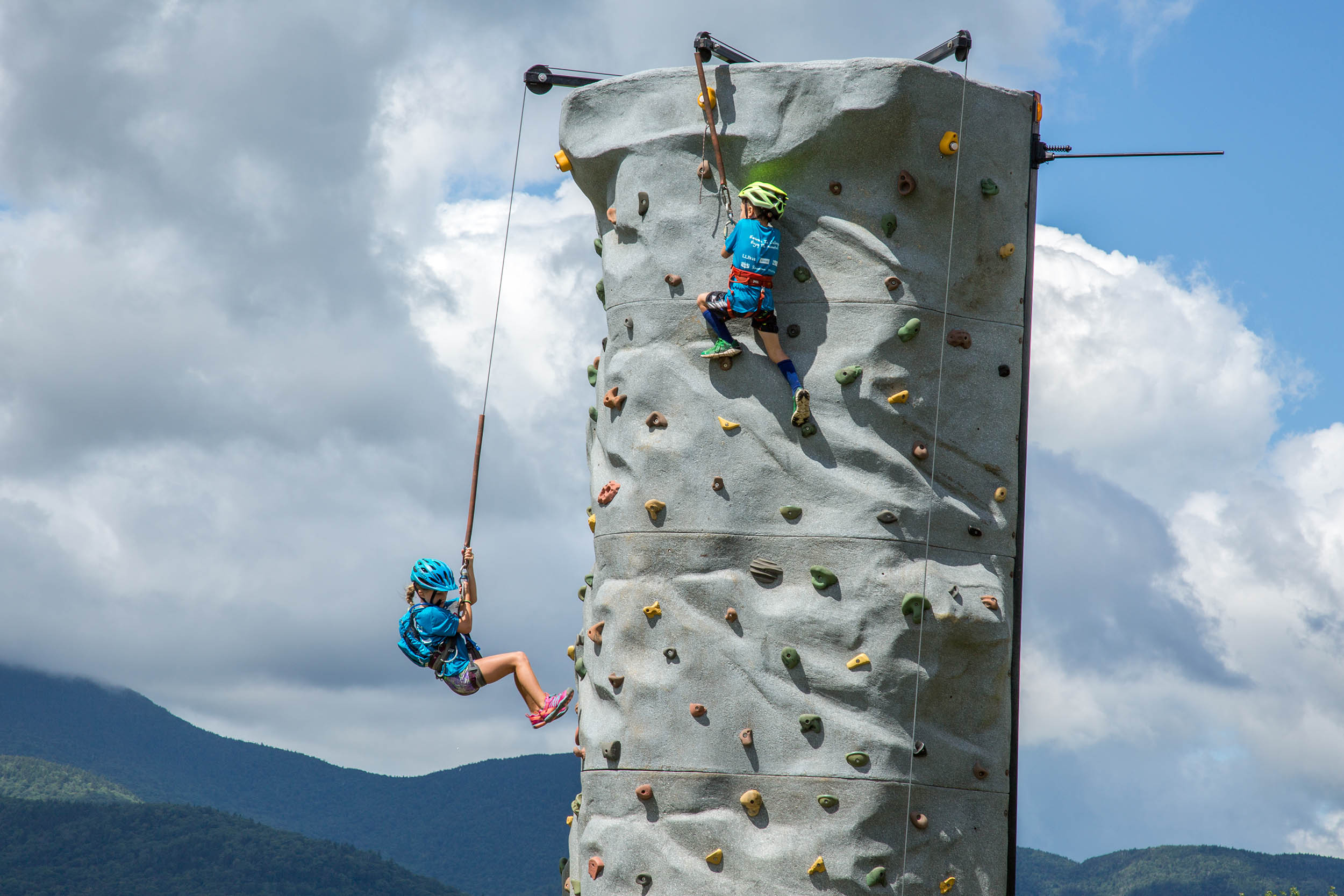 Two children climbing the 24ft mobile rock climbing wall against a mountain backdrop