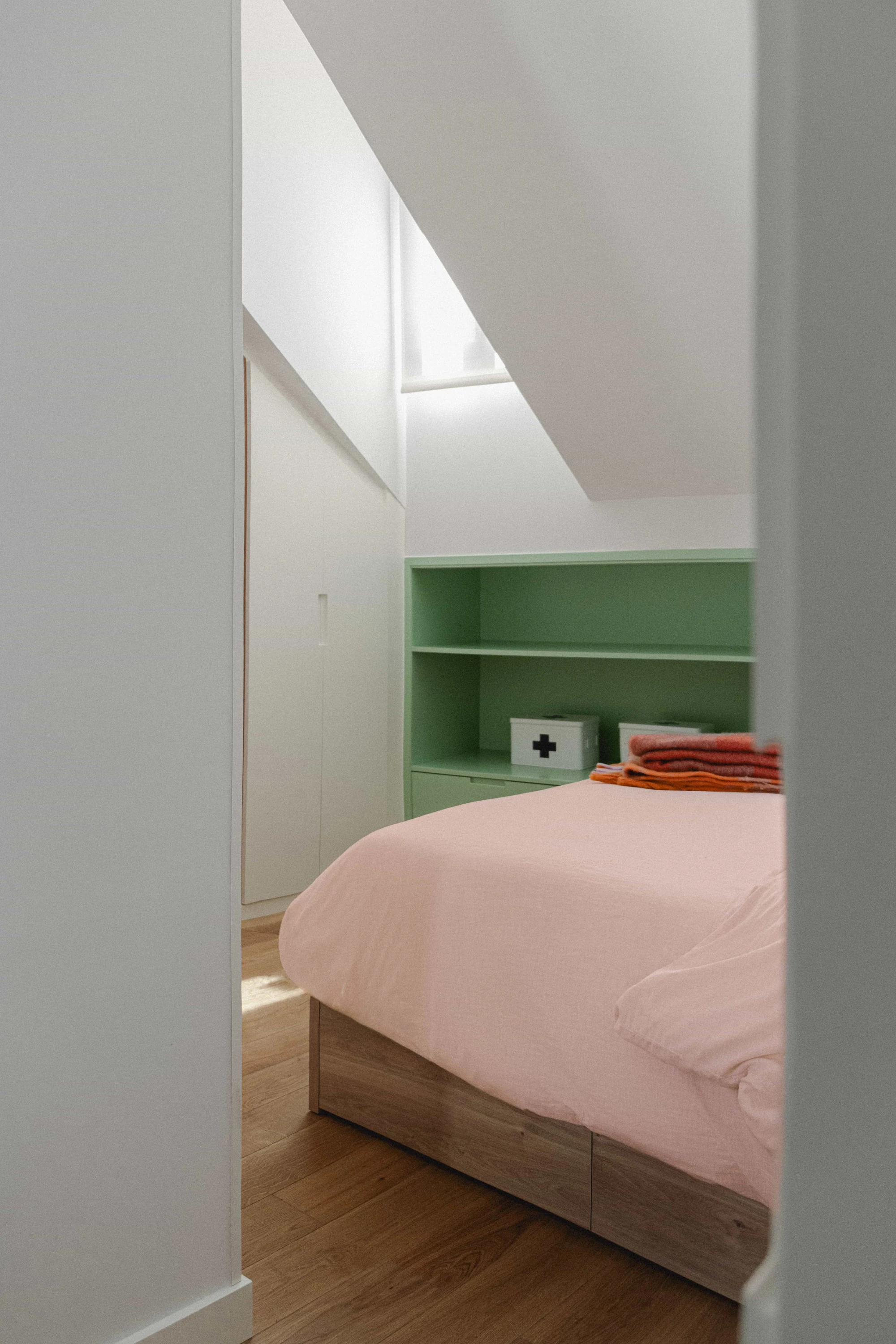 Dormitorio abuhardillado con ropa de cama rosa y mueble verde.