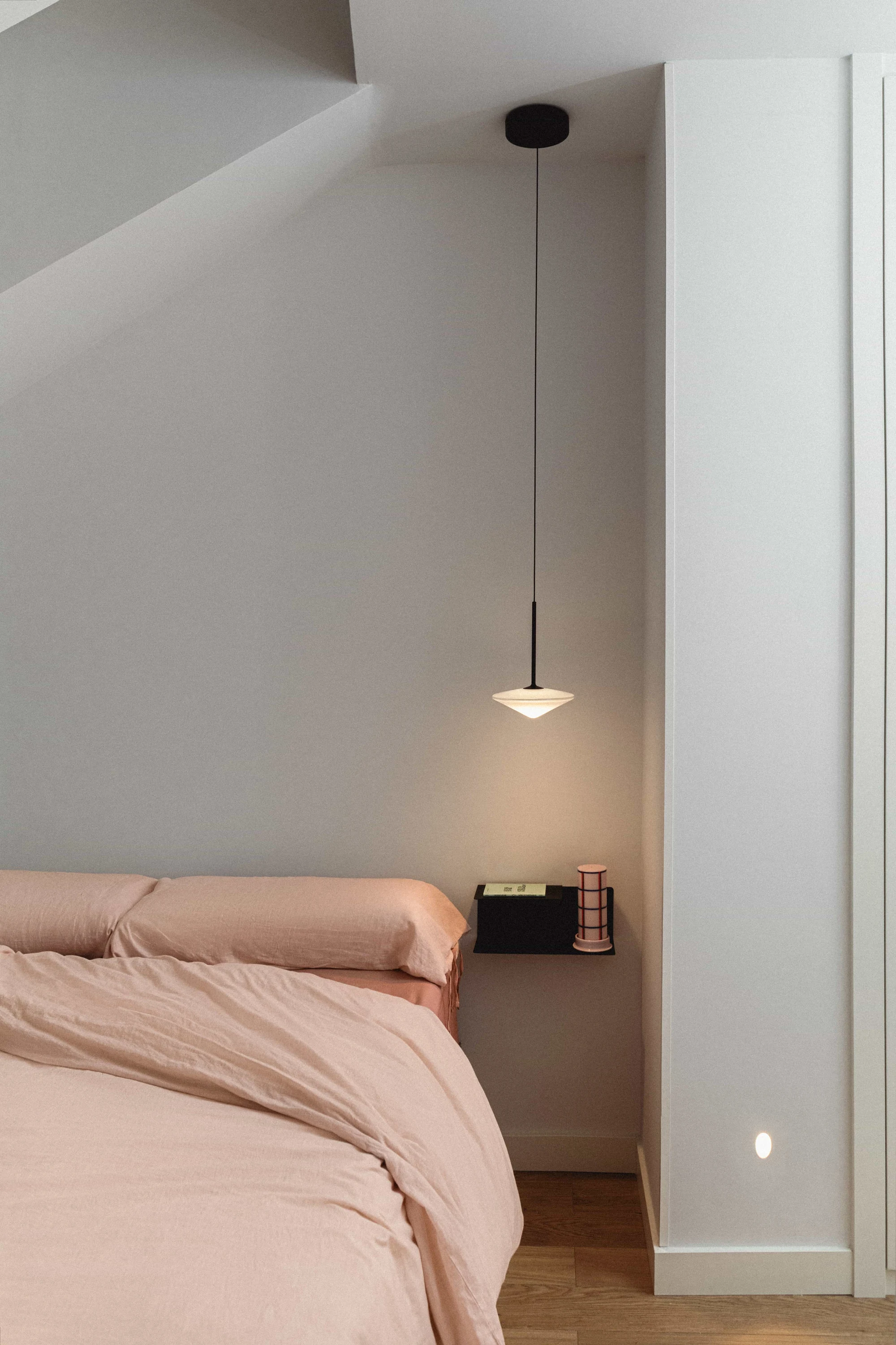 Dormitorio abuhardillado con ropa de cama rosa, lámpara colgante y mesita de noche flotante negra.