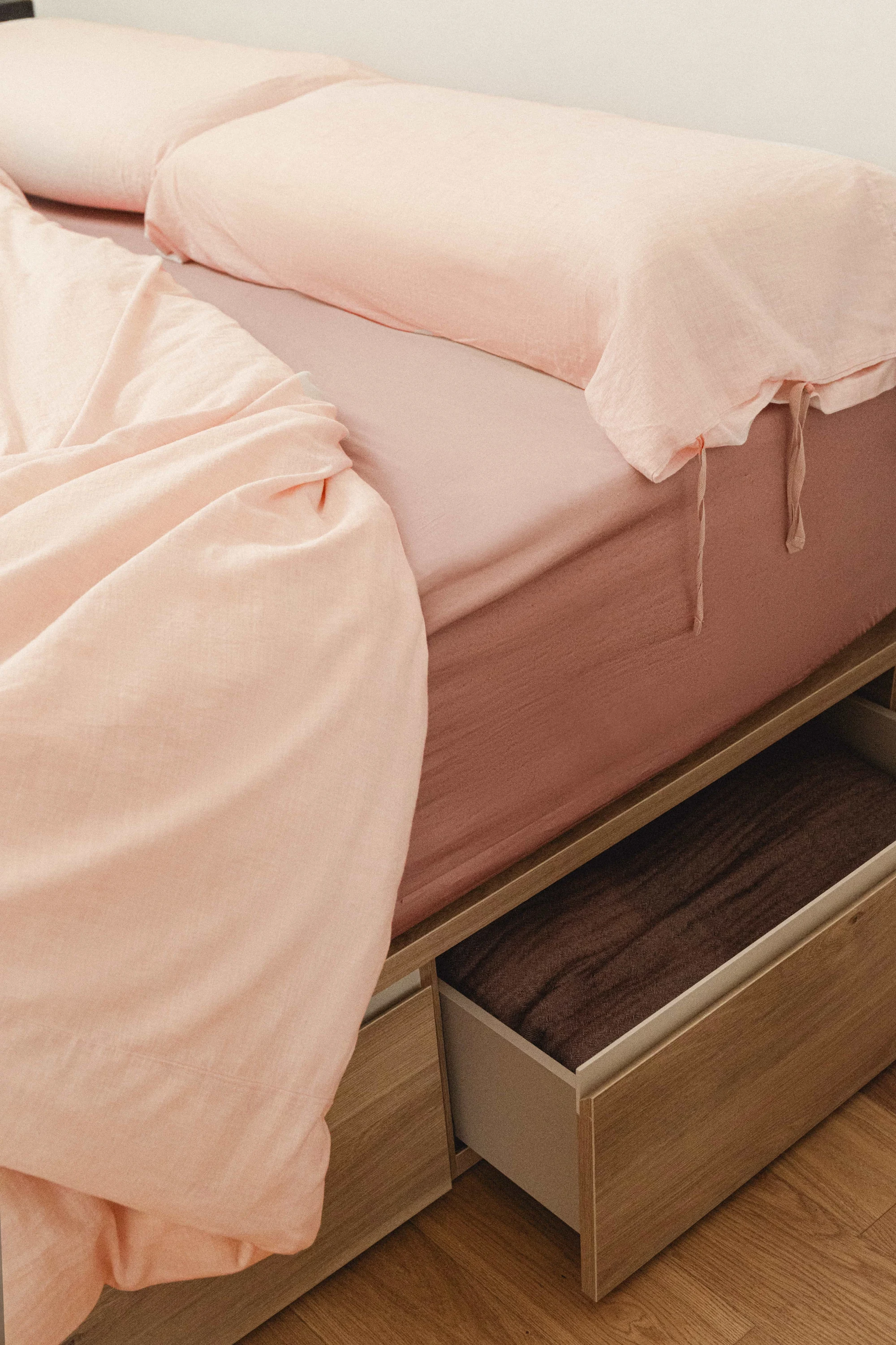 Cama con ropa de cama rosa y canapé de madera con cajón de almacenaje abierto.