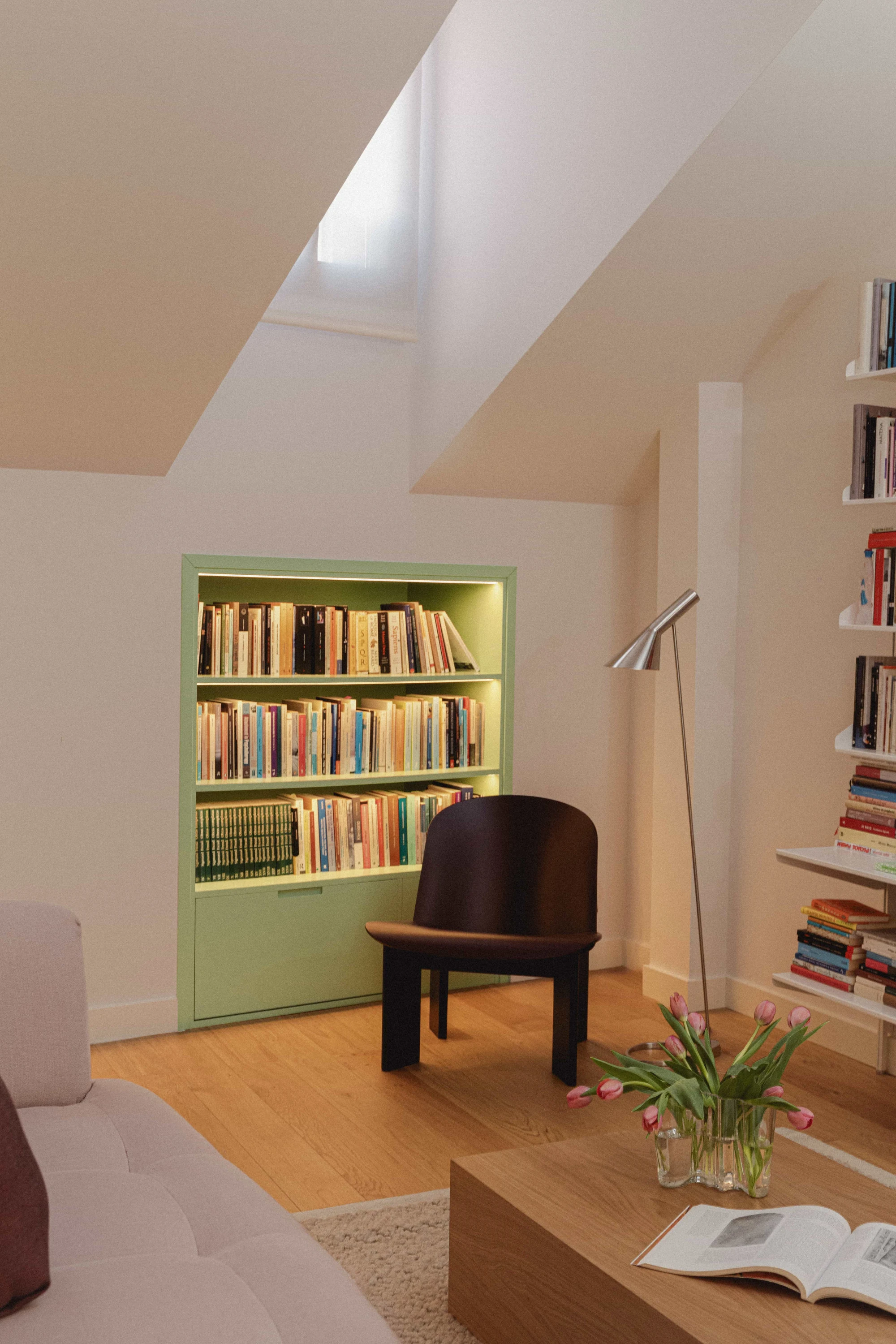 Rincón de lectura abuhardillado con librería empotrada verde menta iluminada, butaca oscura y lámpara de pie.