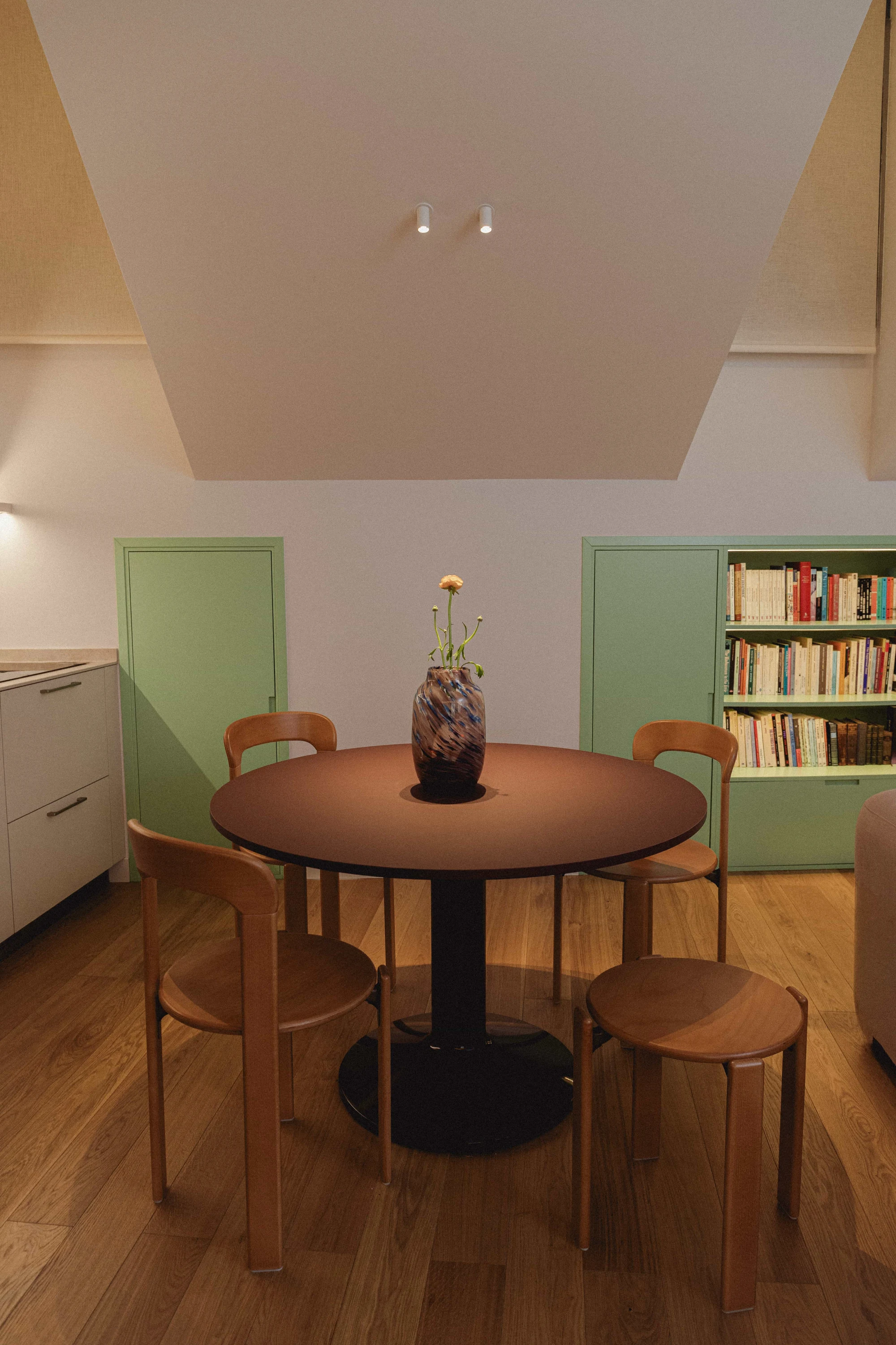 Comedor bajo buhardilla con mesa redonda burdeos, sillas de madera y mueble a medida verde menta con librería.